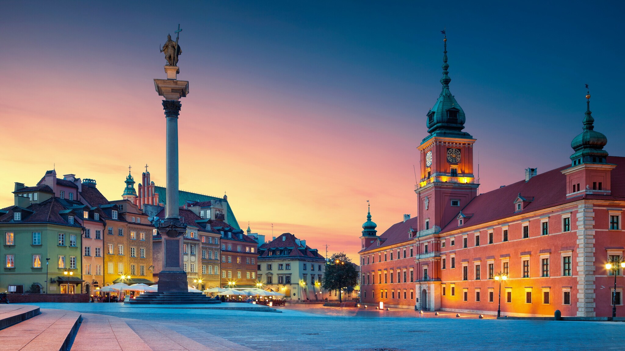 Beleuchteter Marktplatz in der Altstadt von Warschau mit Schlossbau aus rotem Stein und Siegessäule bei Abenddämmerung.