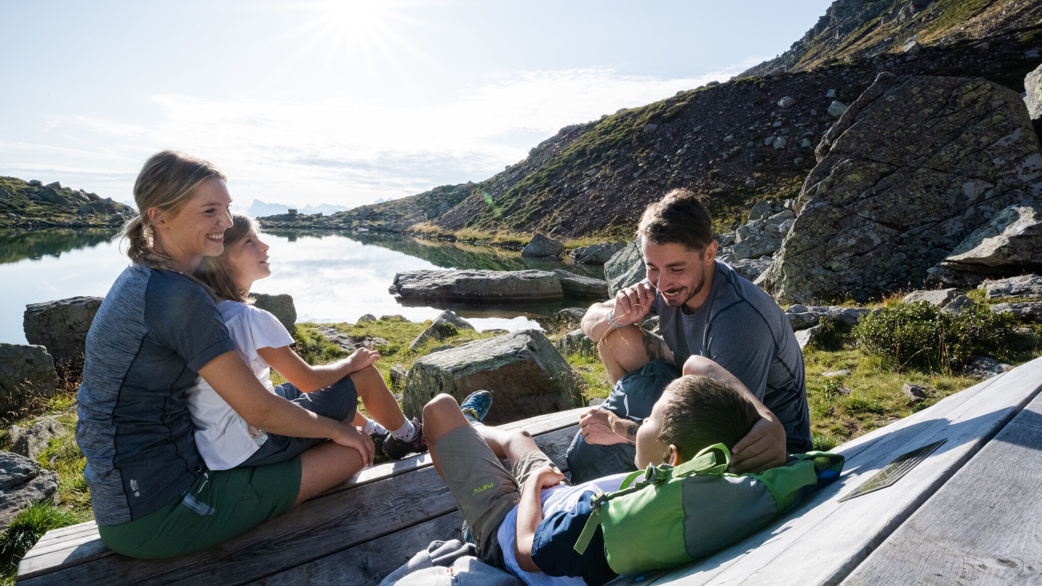 Eine Familie mit zwei Kindern in Wanderkleidung sitzt auf einer Holzbank an einem See in den Bergen. Eine Familie mit zwei Kindern in Wanderkleidung sitzt auf einer Holzbank an einem See in den Bergen.