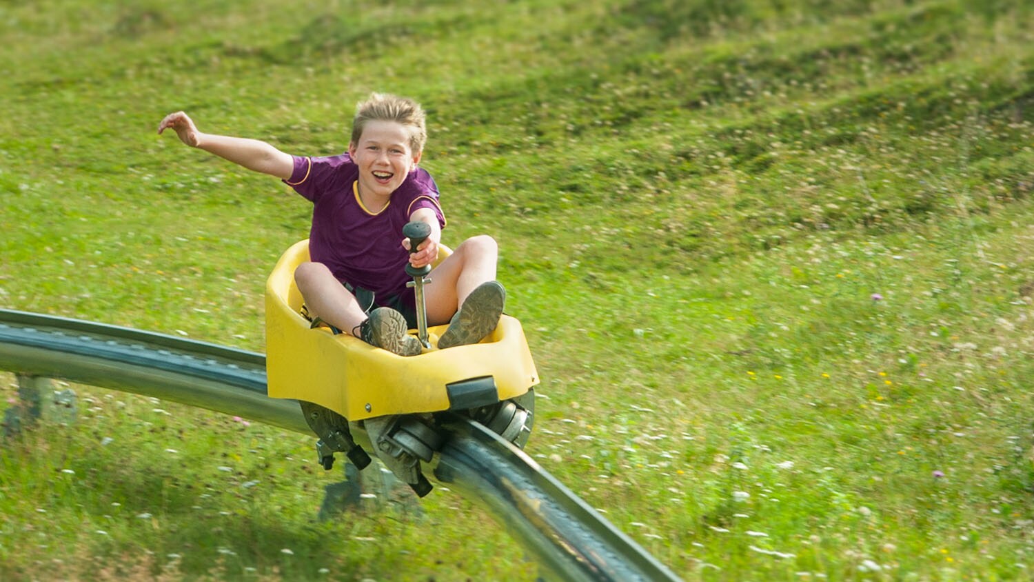 Ein fröhlicher Junge fährt in einem Bobschlitten auf einer Schiene über eine grüne Wiese. Ein fröhlicher Junge fährt in einem Bobschlitten auf einer Schiene über eine grüne Wiese.