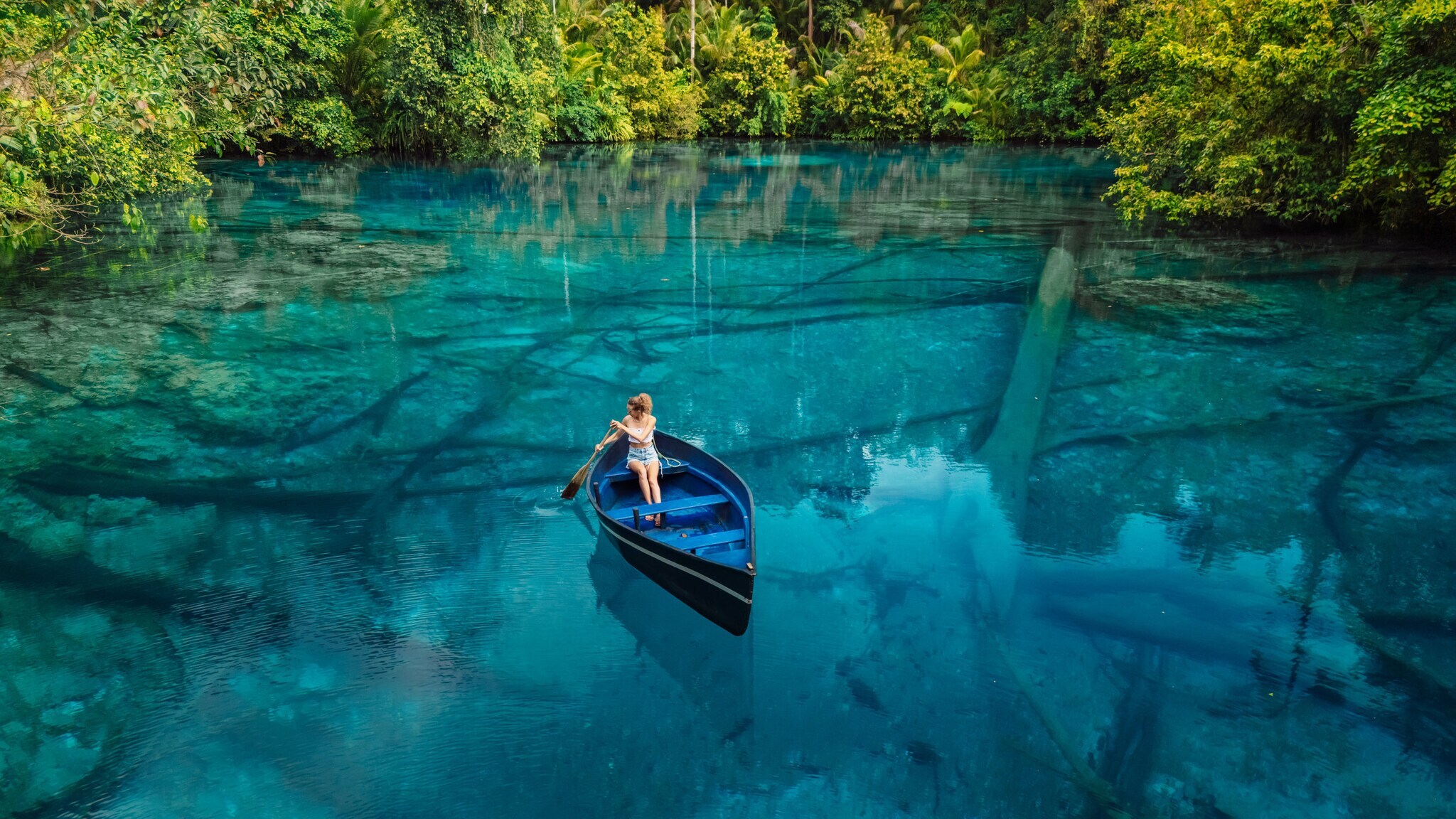 Eine Frau sitzt in einem Ruderboot auf einem blauen, glasklaren See, umgeben von grüner Vegetation.