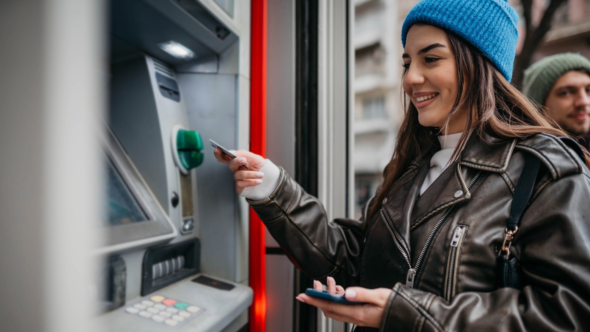 Eine Frau mit Mütze zieht mit einer Kreditkarte Geld an einem Bankautomaten auf der Straße.