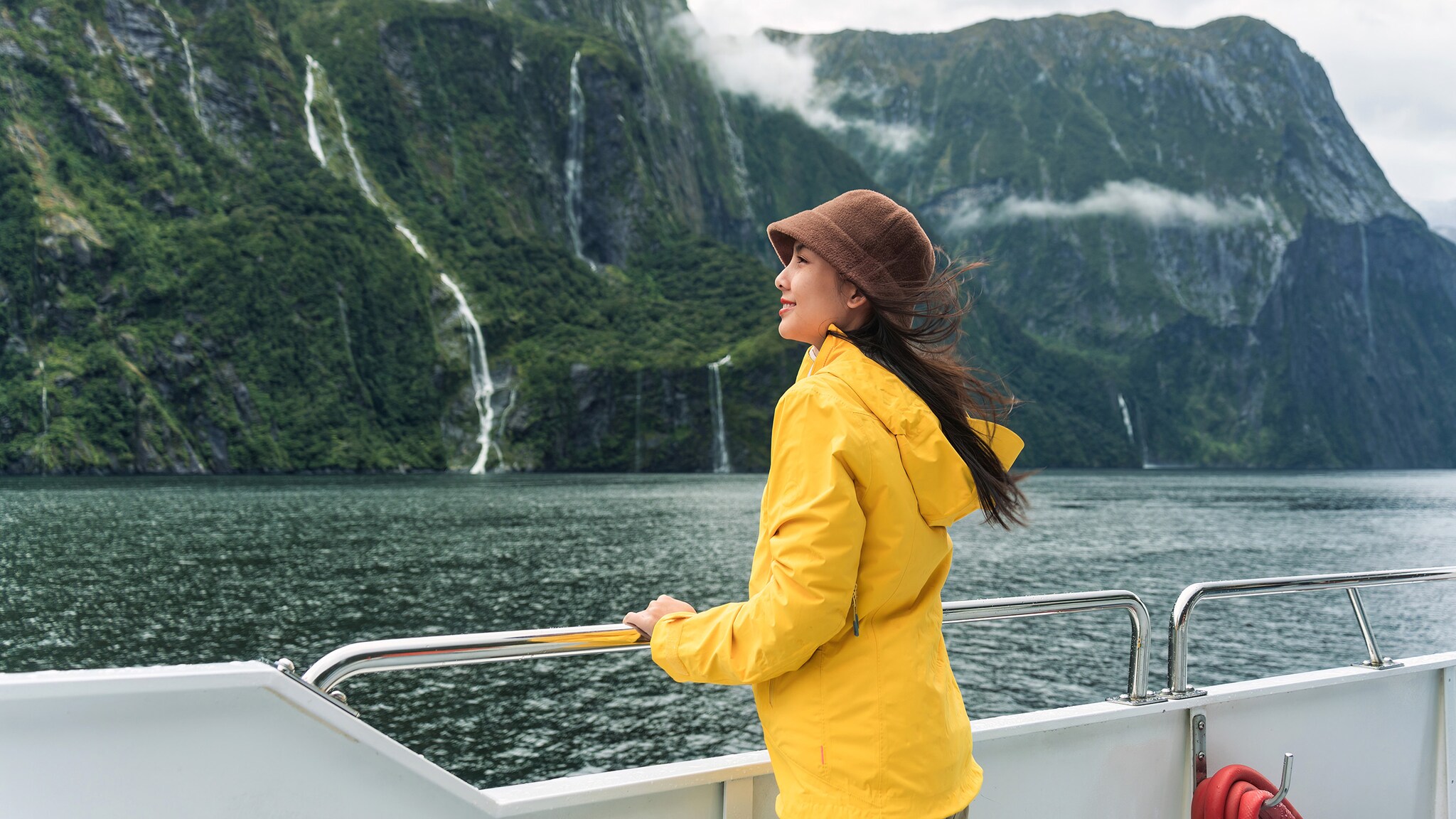 Eine Frau im gelben Regenmantel steht an der Reling eines Schiffes in einer Fjordlandschaft.