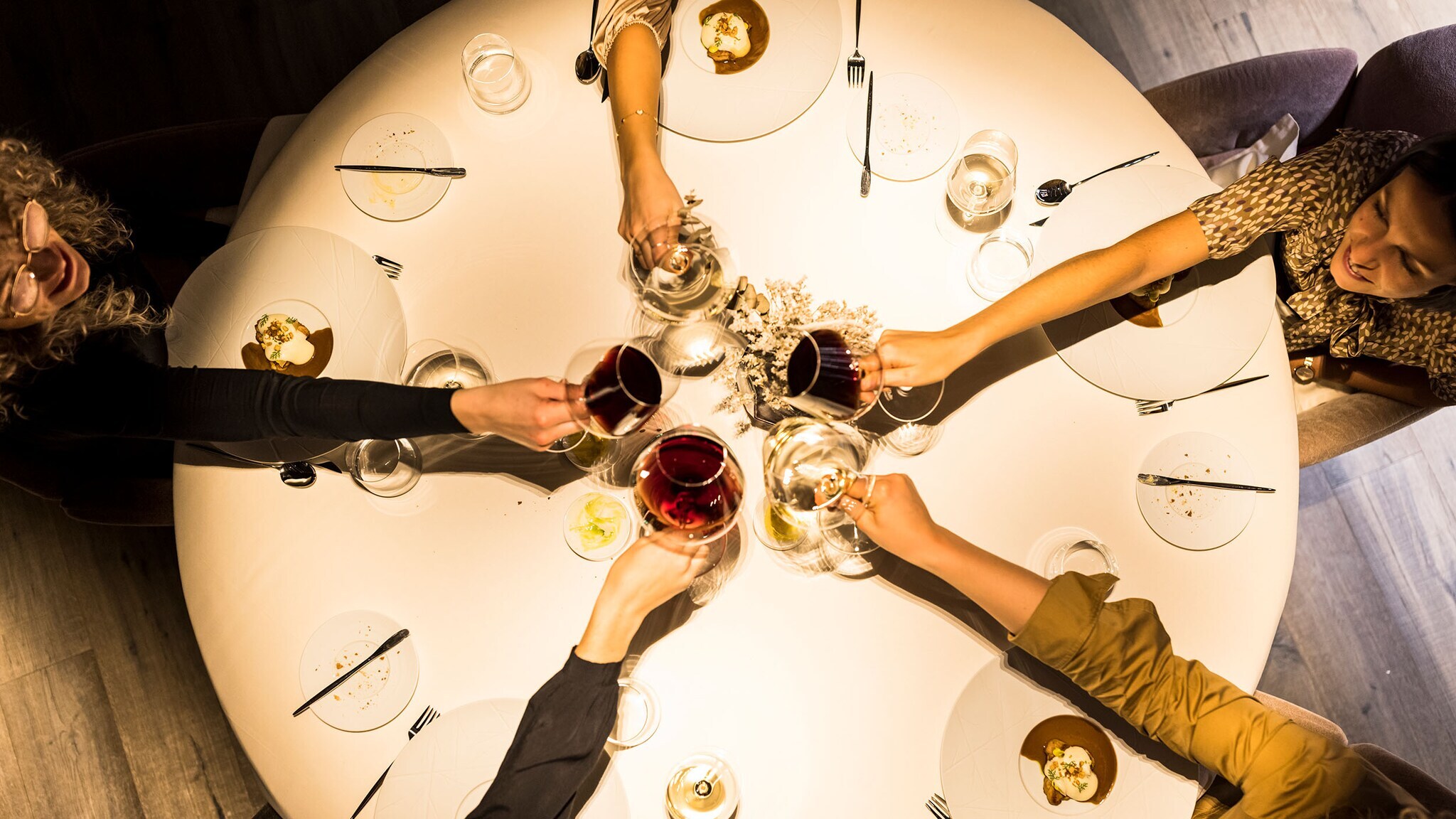 Aufsicht von fünf Armen mit Weingläsern beim Anstoßen über einem runden Esstisch bei dämmrigen Licht.
