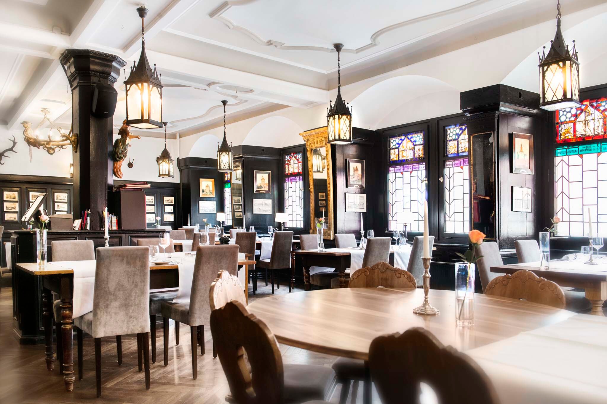Urig-gemütlicher Innenraum des Restaurants Weinstube mit Buntglasfenstern, edlen Wandvertäfelungen, Rundbögen, verzierter Decke sowie historischen Lampen und Bildern an den Wänden.