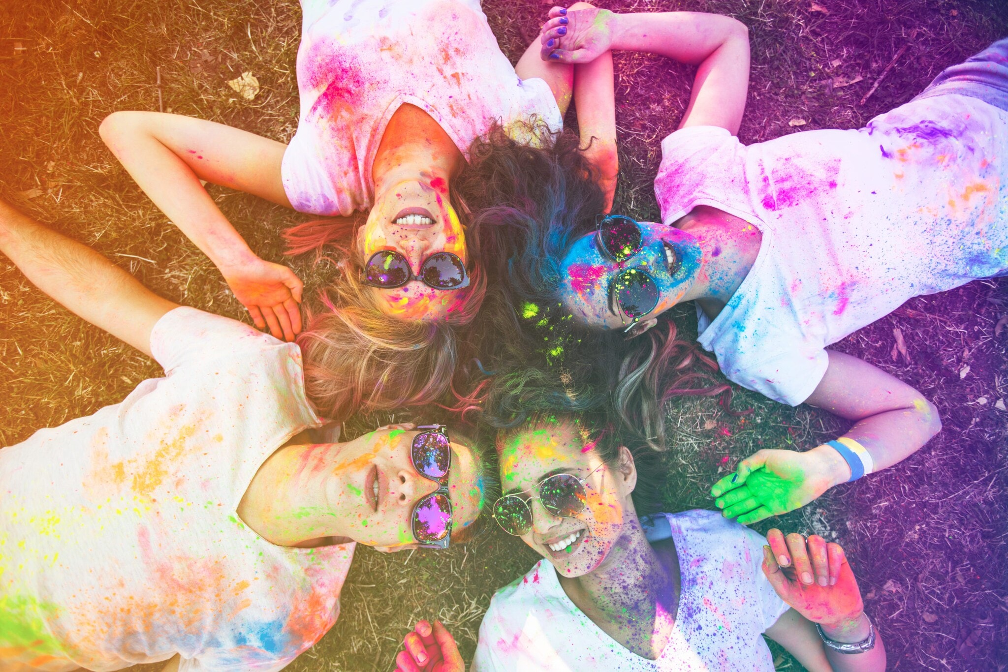 Vier Festivalbesucherinnen mit Sonnenbrillen und farbig besprenkelten Shirts, Gesichtern und Händen liegen lachend auf dem Rasen. Vier Festivalbesucherinnen mit Sonnenbrillen und farbig besprenkelten Shirts, Gesichtern und Händen liegen lachend auf dem Rasen.