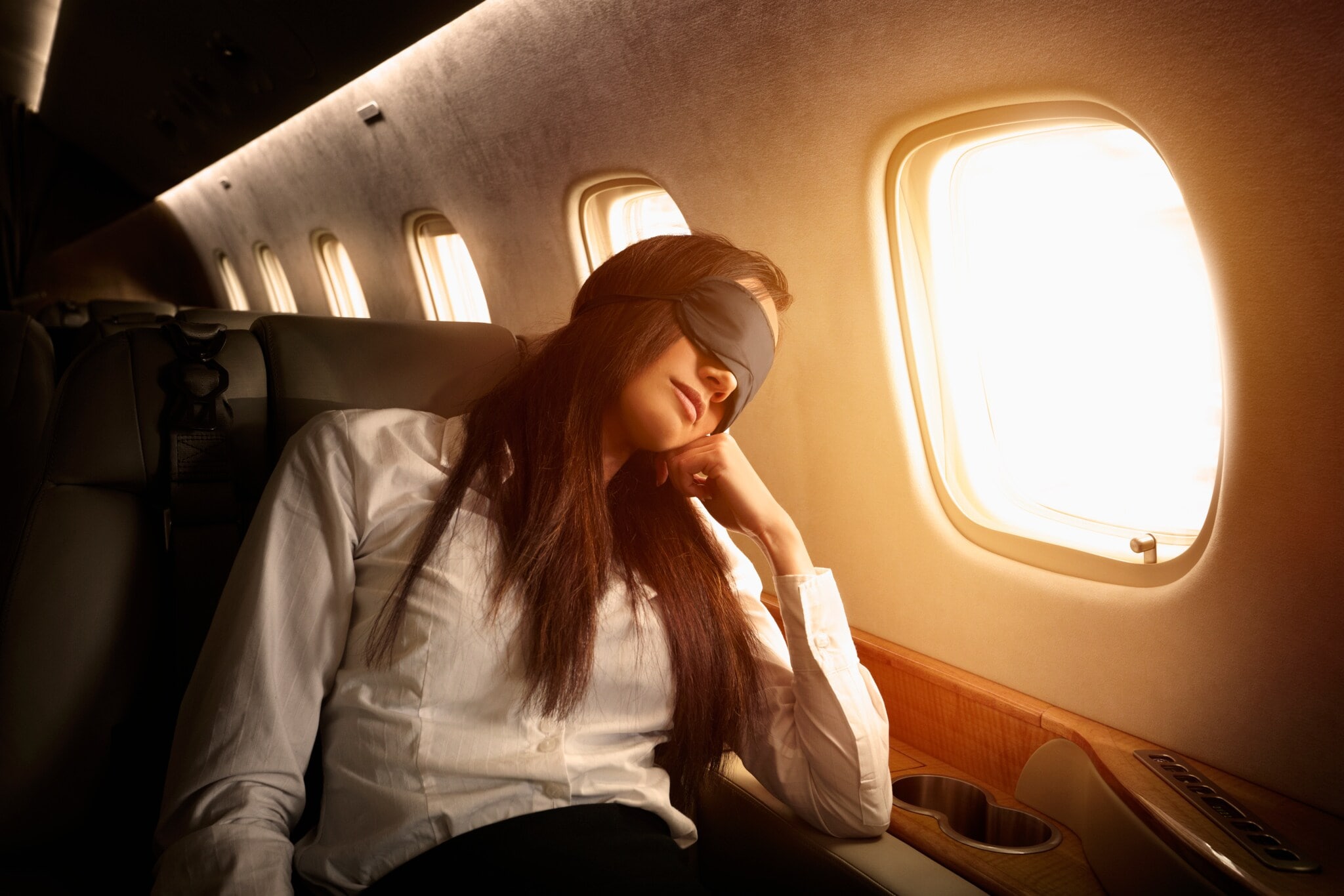 Eine Frau mit Schlafmaske stützt ihren Kopf auf dem Arm an einem Fenster in der Business Class eines Flugzeuges. Eine Frau mit Schlafmaske stützt ihren Kopf auf dem Arm an einem Fenster in der Business Class eines Flugzeuges.