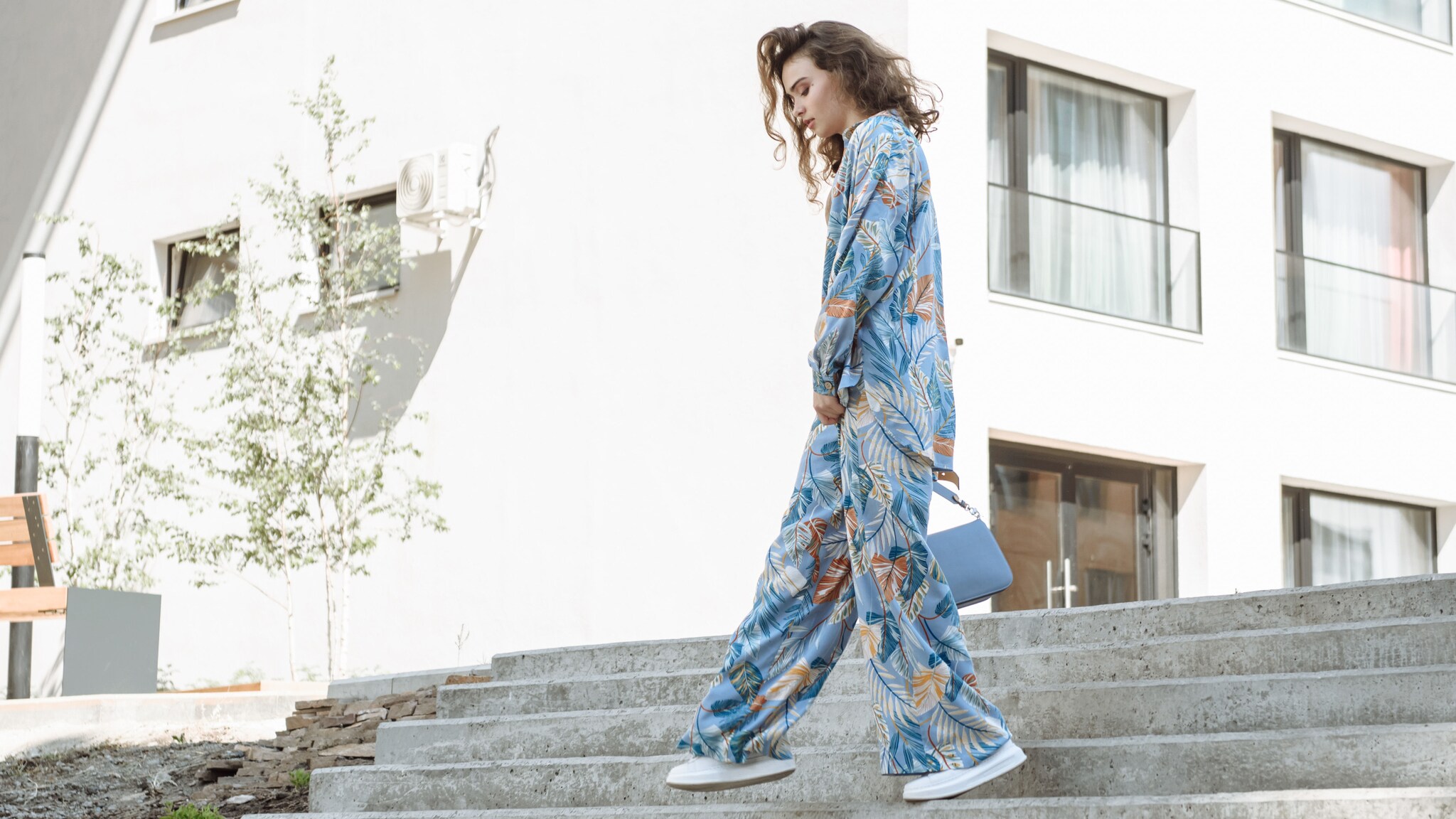 Junge Frau in blauem Blumen-Pyjama, weißen Turnschuhen und einer schicken blauen Tasche geht eine Betontreppe hinunter
