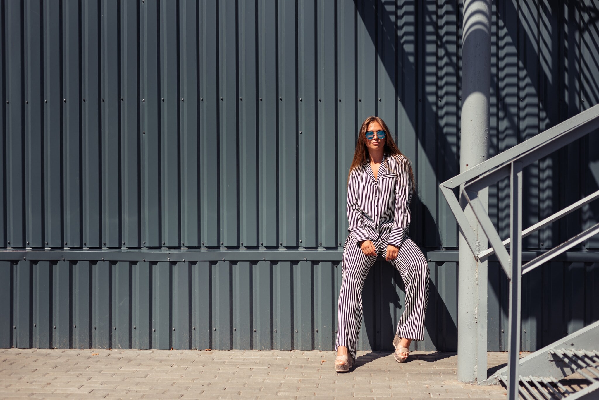 Junge Frau mit Sonnenbrille in blau-weiß gestreiftem Pyjama lehnt an eine Wand aus grauem festem Wellblech. Junge Frau mit Sonnenbrille in blau-weiß gestreiftem Pyjama lehnt an eine Wand aus grauem festem Wellblech.