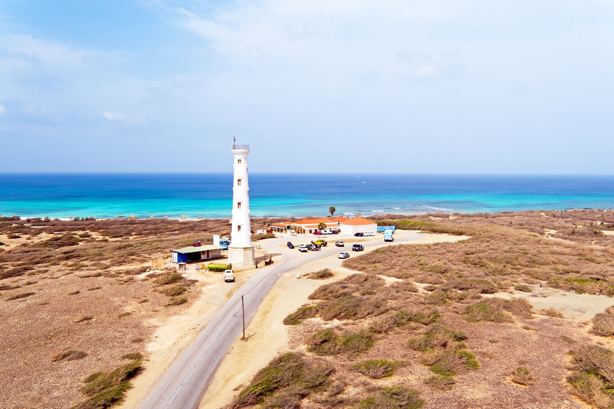 Blick von oben auf einen weißen Leuchtturm in Aruba, im Hintergrund türkisblaues Meer.