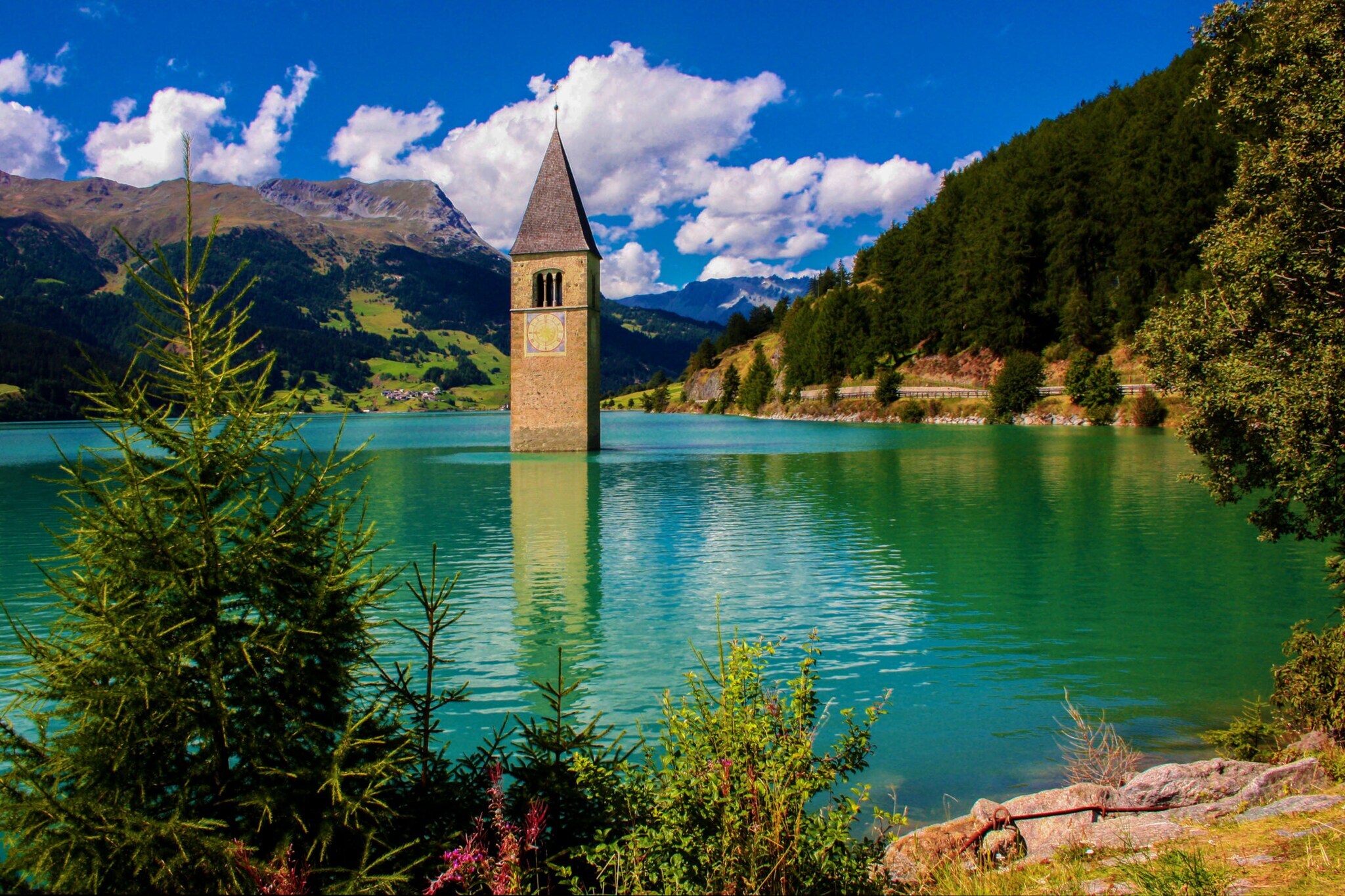 Blick auf den Reschensee, ein malerischer Stausee mit aus dem Wasser ragenden Kirchturm, umgeben von grüner Landschaft.