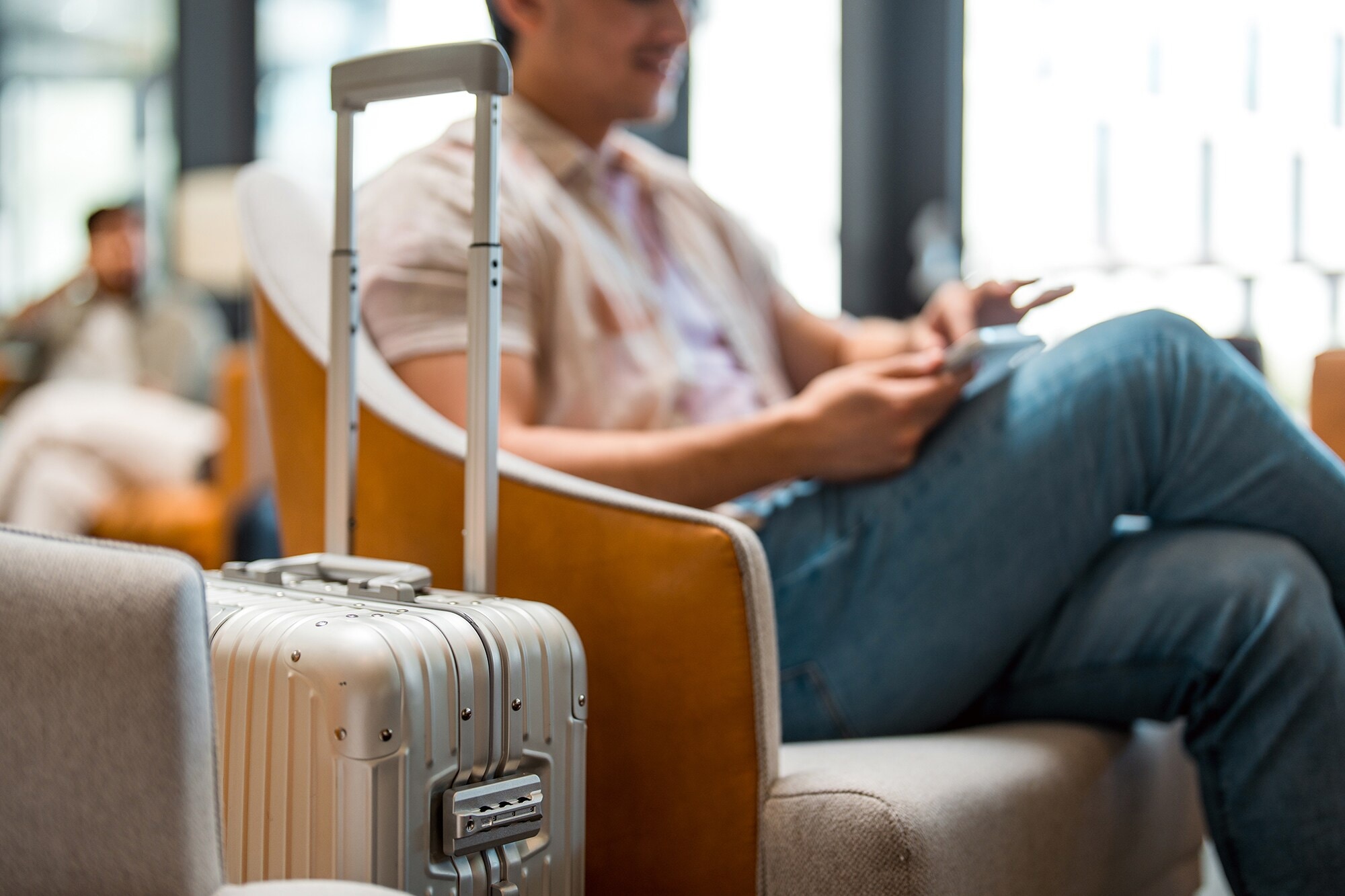 Packliste für deine Geschäftsreise: Smarte Tipps und nützliche Gadgets Ein Mann sitzt im Wartebereich eines Flughafens. Im Vordergrund steht sein Rollkoffer.