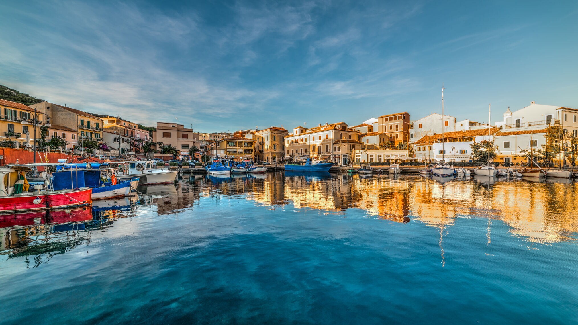 Hafen mit bunten Booten und Gebäuden am Ufer unter bewölktem Himmel, Spiegelung im Wasser.