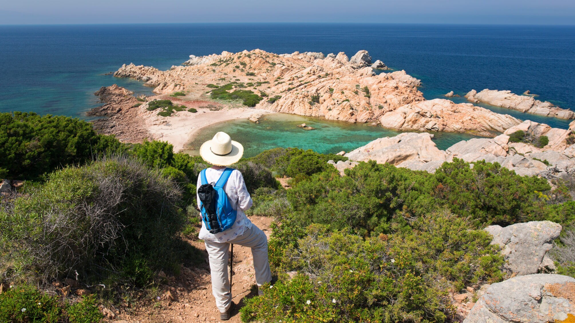Wandernde Person mit weißem Hut und blauem Rucksack blickt auf felsige Küste und türkisfarbenes Meer.