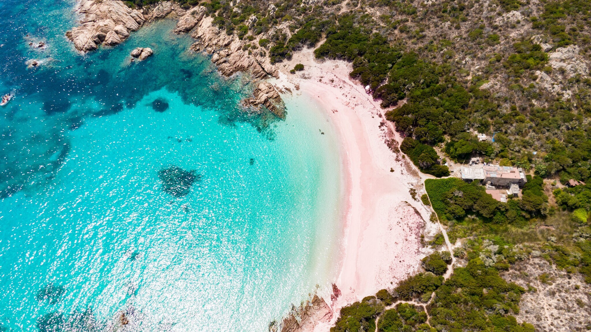 Luftaufnahme einer abgelegenen Bucht mit türkisfarbenem Wasser, rosafarbenen Sandstrand und umgeben von dichtem Grün und Felsen.