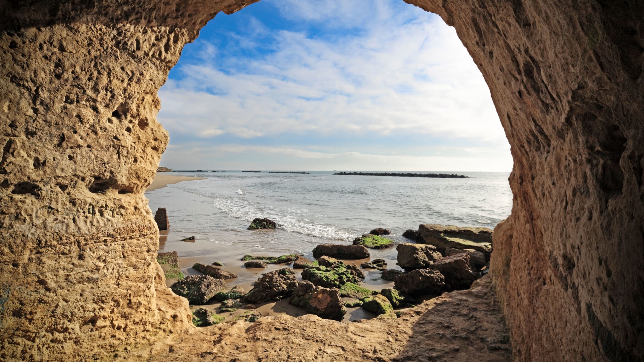 Blick auf einen Sandstrand mit kleinen Felsen im Meer durch eine runde Öffnung in einem großen Felsen am Strand.