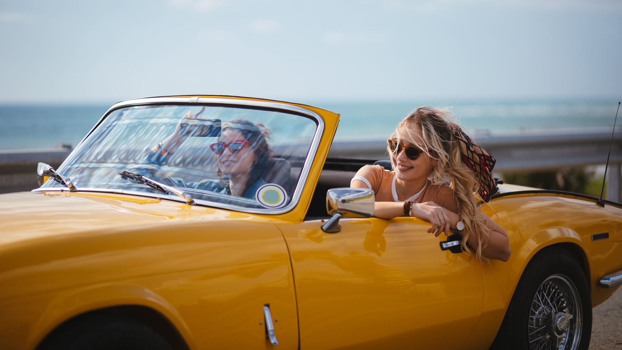 Zwei junge Frauen mit Sonnenbrillen in einem gelben Cabriolet Oldtimer auf einer Küstenstraße.