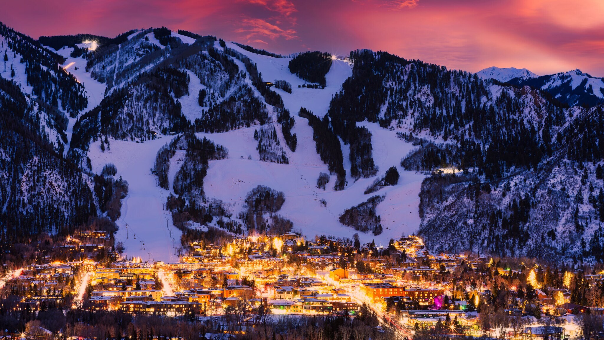 Bergdorf mit beleuchteten Gebäuden und schneebedeckten Skipisten bei Sonnenuntergang.