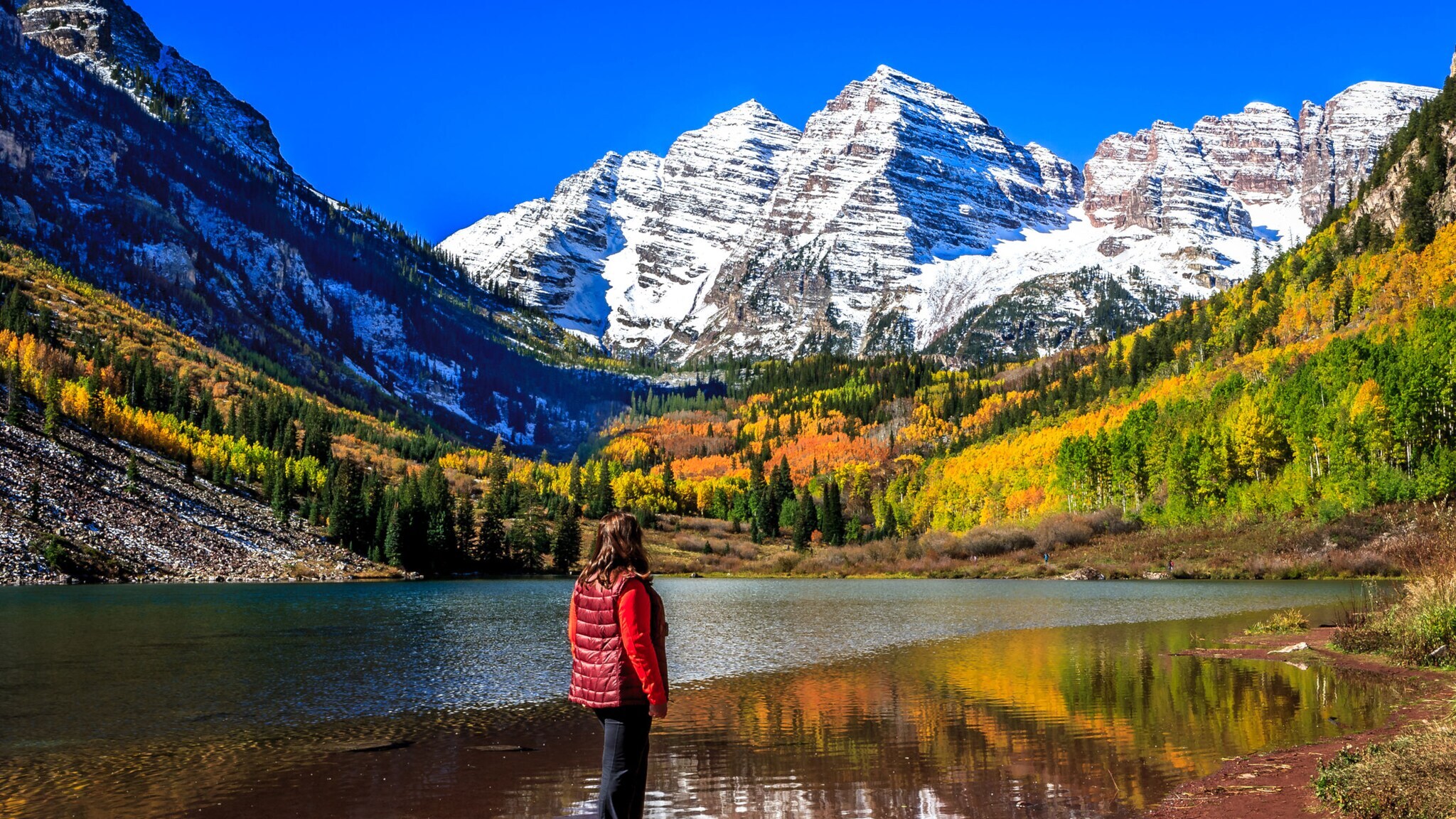 Person in roter Weste steht am Ufer eines Sees mit Blick auf schneebedeckte Berge und herbstlich gefärbten Wald.