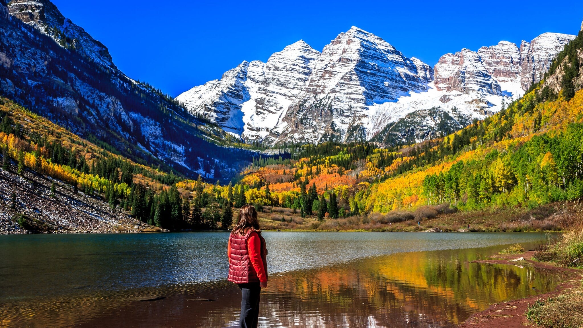 Person in roter Weste steht am Ufer eines Sees mit Blick auf schneebedeckte Berge und herbstlich gefärbten Wald. Person in roter Weste steht am Ufer eines Sees mit Blick auf schneebedeckte Berge und herbstlich gefärbten Wald.