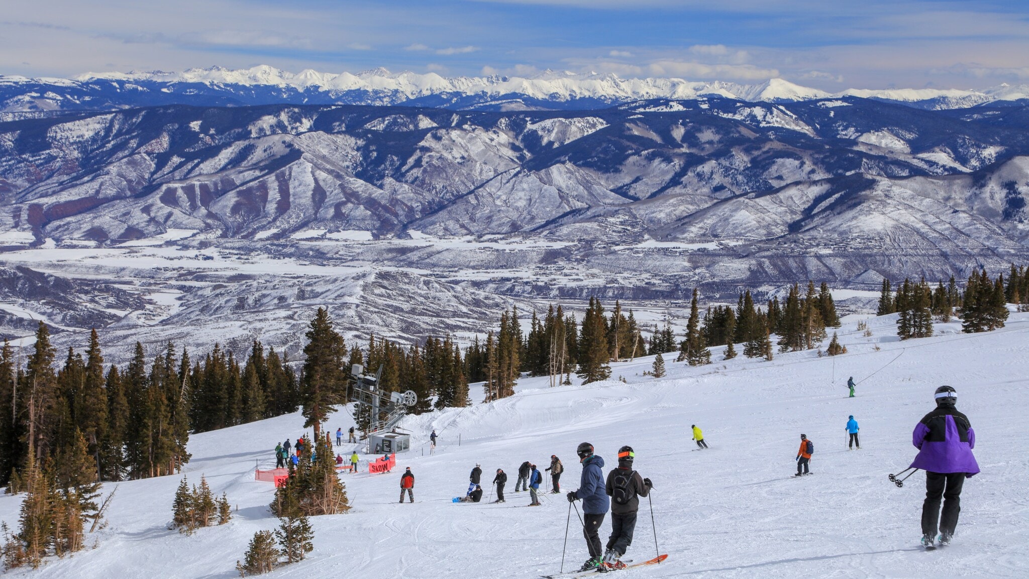 Skifahrende Personen auf schneebedecktem Hang mit Tannenbäumen und Bergen im Hintergrund unter bewölktem Himmel. Skifahrende Personen auf schneebedecktem Hang mit Tannenbäumen und Bergen im Hintergrund unter bewölktem Himmel.
