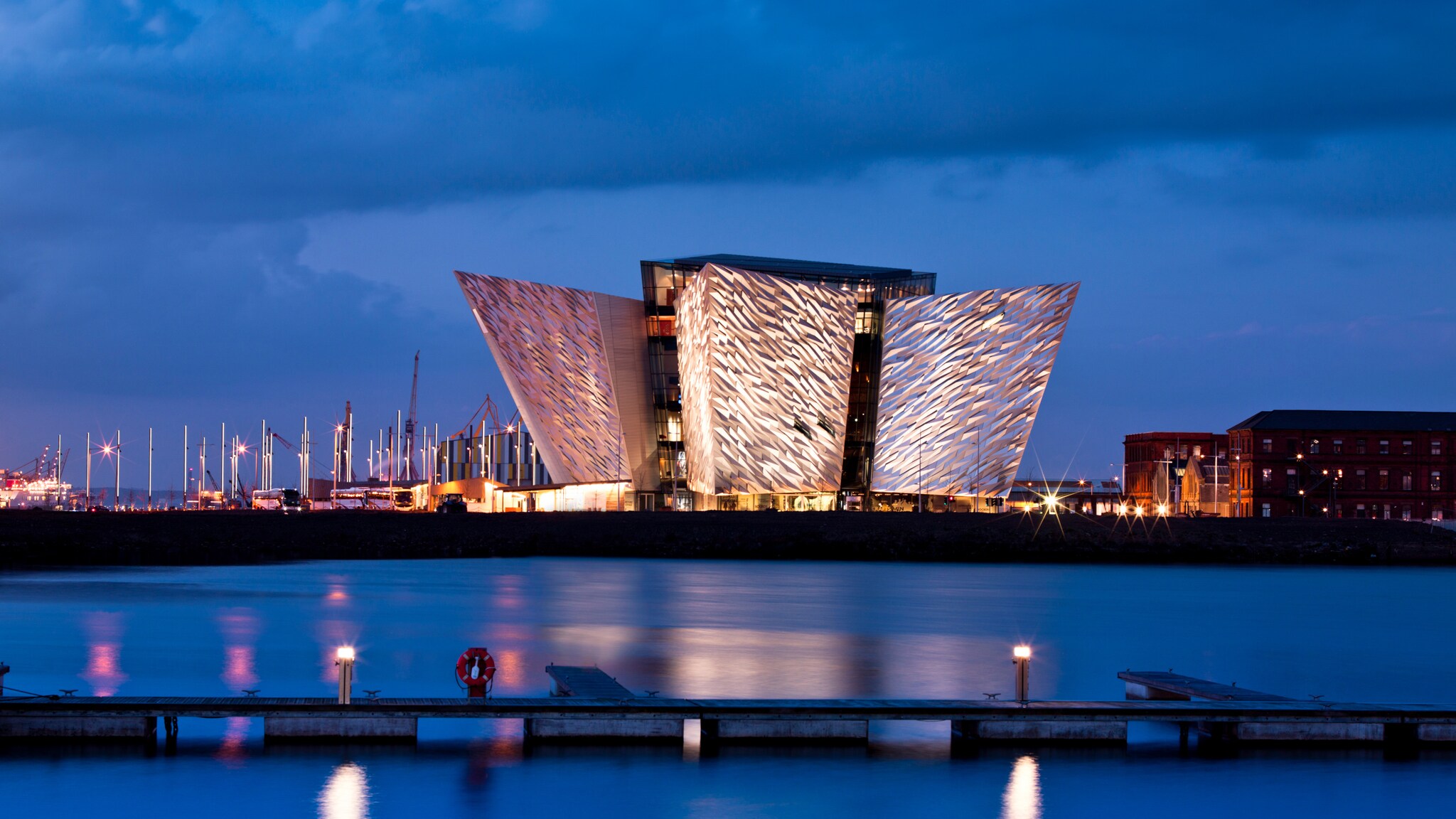 Beleuchtetes modernes Gebäude mit markanter, gefalteter Fassade am Wasser bei Abenddämmerung Beleuchtetes modernes Gebäude mit markanter, gefalteter Fassade am Wasser bei Abenddämmerung