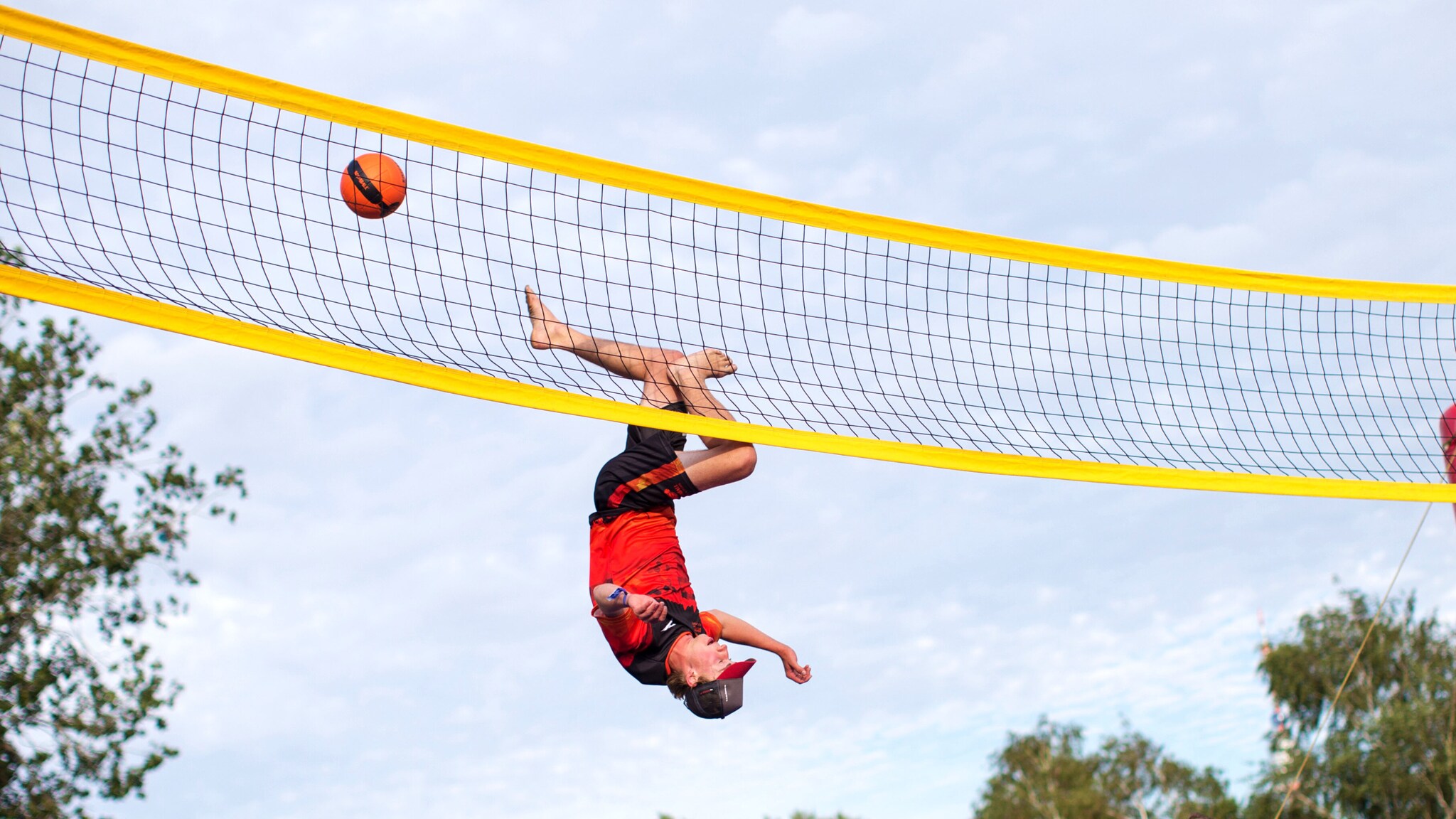 Ein Sportler kopfüber bei einem Sprung an einem Volleyballnetz, daneben ein fliegender Ball. Ein Sportler kopfüber bei einem Sprung an einem Volleyballnetz, daneben ein fliegender Ball.