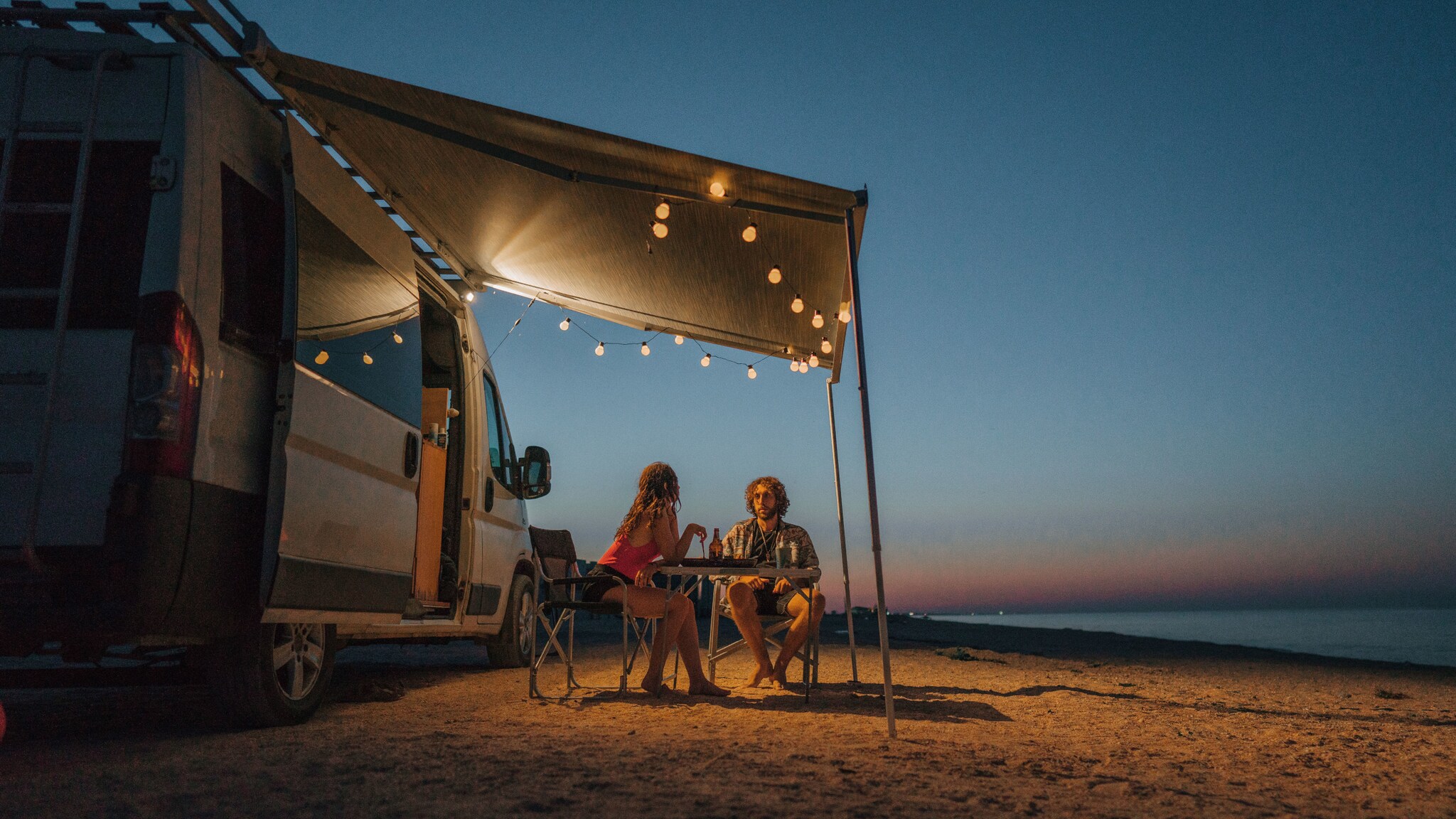 Ein Paar sitzt an einem Campingtisch unter der beleuchteten Markise eines Wohnmobils an einem Sandstrand bei Dunkelheit.