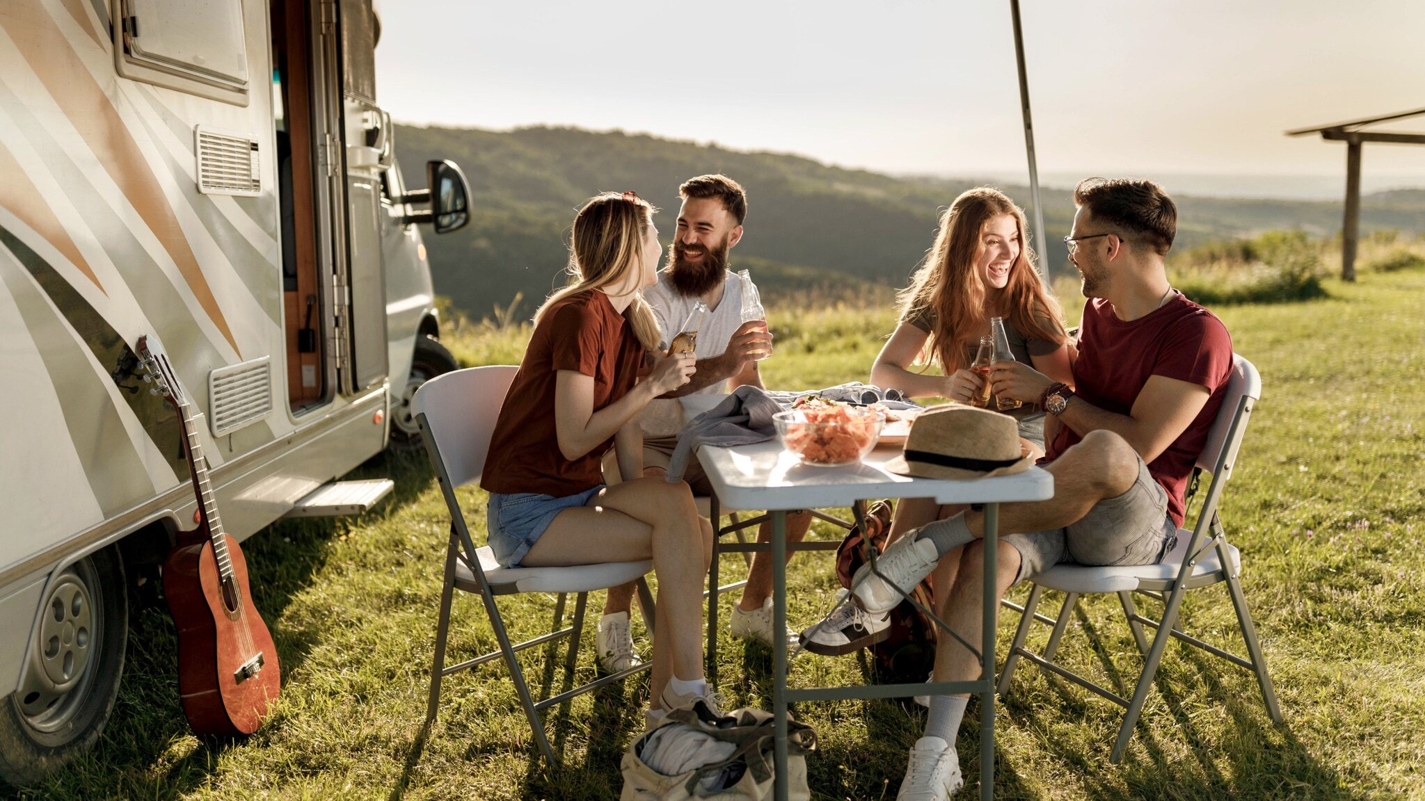 Vier gut gelaunte Personen in Sommerkleidung sitzen an einem Campingtisch neben einem Wohnmobil auf einer Rasenfläche vor Hügellandschaft. Vier gut gelaunte Personen in Sommerkleidung sitzen an einem Campingtisch neben einem Wohnmobil auf einer Rasenfläche vor Hügellandschaft.