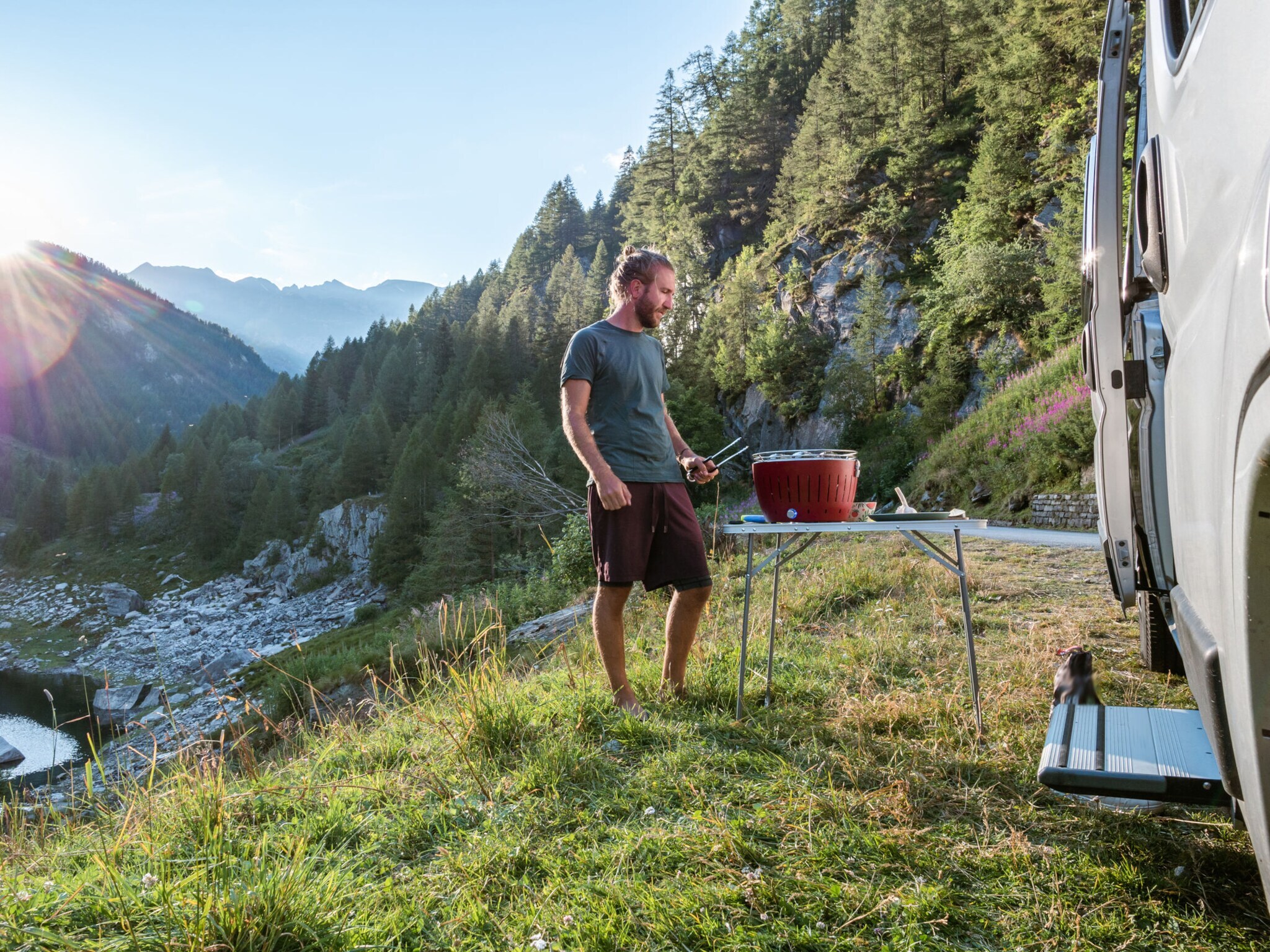 Ein Mann steht mit einer Grillzange an einem Tisch mit tragbarem Grill neben einem Wohnmobil in einer Berglandschaft. Ein Mann steht mit einer Grillzange an einem Tisch mit tragbarem Grill neben einem Wohnmobil in einer Berglandschaft.