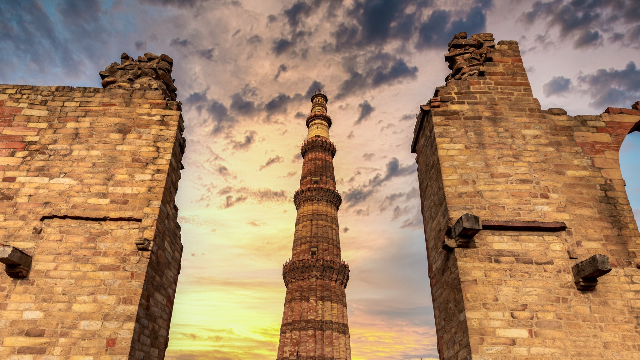 Das Qutub Minar, ein hoher roter Sandsteinturm, umgeben von Ruinenmauern bei Sonnenuntergang mit bewölktem Himmel