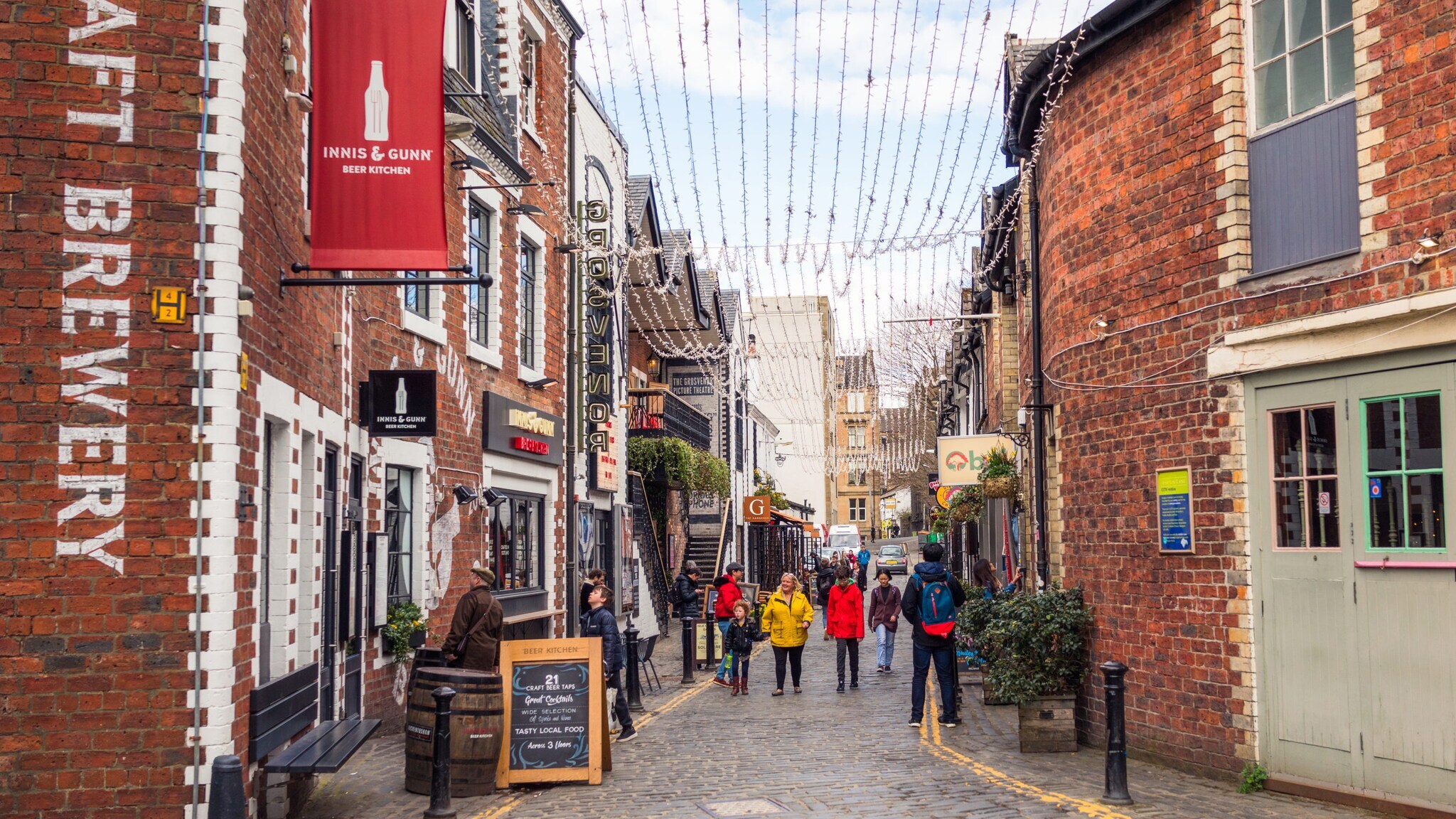Belebte Kopfsteinpflasterstraße mit Geschäften und Restaurants, darunter eine Craft Brewery, Menschen gehen entlang der Straße unter Lichterketten