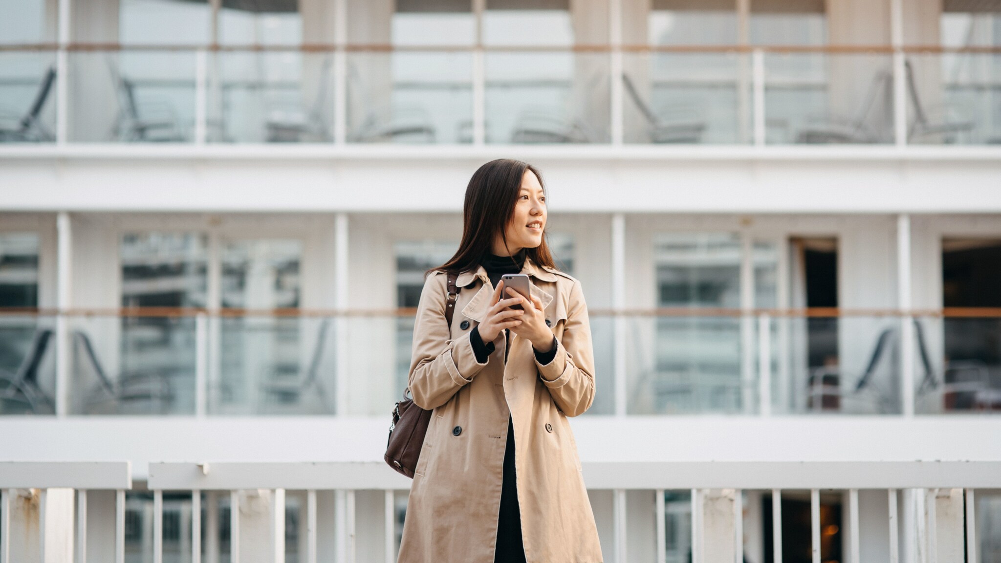 Eine Frau im Mantel steht mit ihrem Smartphone in den Händen vor einem Kreuzfahrtschiff. Eine Frau im Mantel steht mit ihrem Smartphone in den Händen vor einem Kreuzfahrtschiff.
