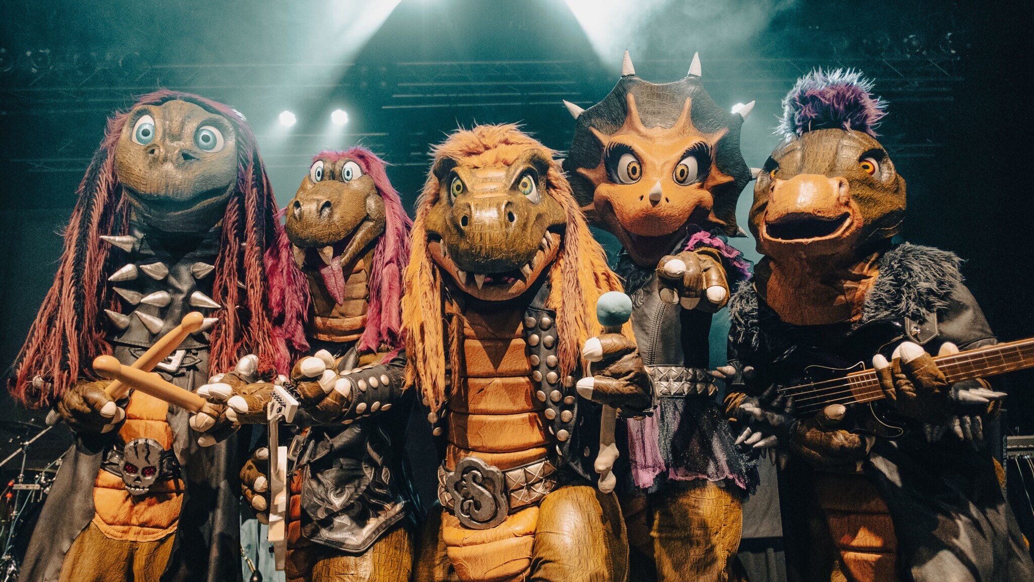 Vier Personen in Dinosaurierkostümen, die in die Kamera gucken und teilweise Instrumente und ein Mikrofon halten