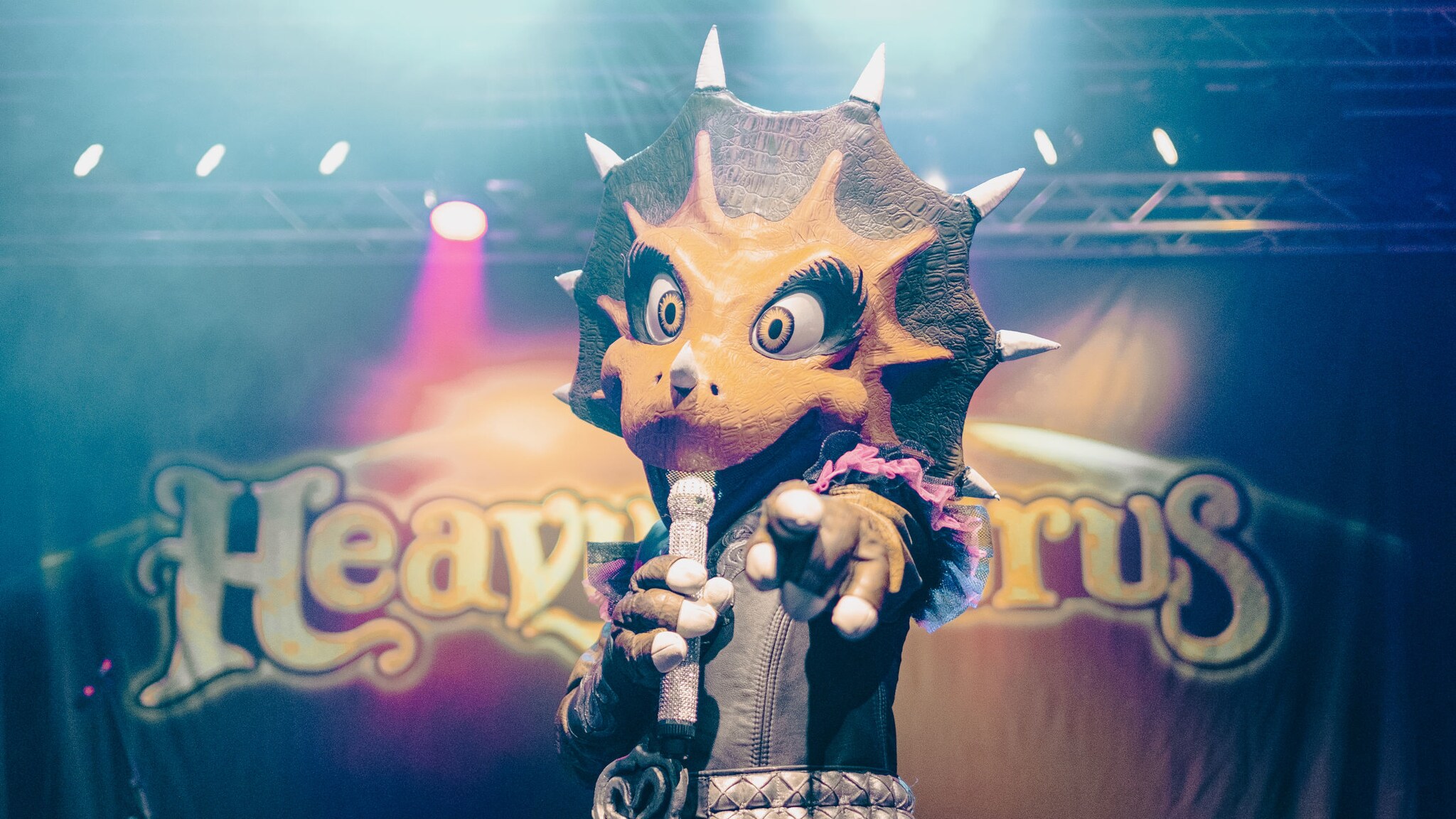 ine Person im Dinosaurierkostüm, die ein Mikrofon hält und mit einer Hand auf die Kamera zeigt, im Hintergrund Bühnenlicht und der Schriftzug „Heavysaurus“