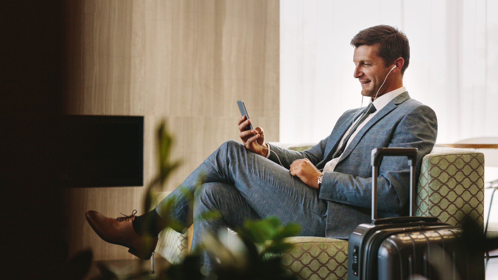 Ein Geschäftsmann sitzt in einem Sessel in einer Flughafen-Lounge und schaut lächelnd auf sein Smartphone.