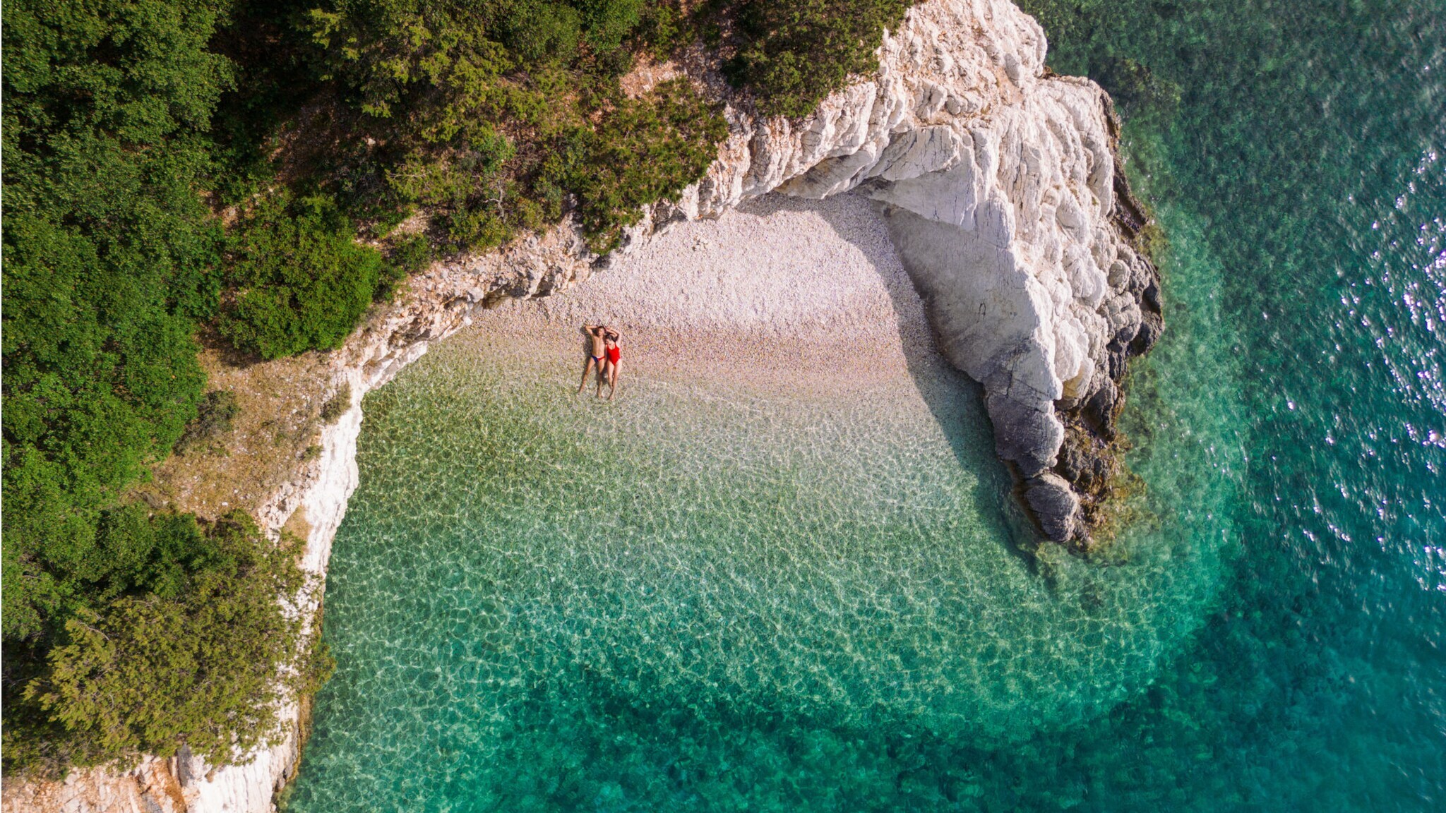 Luftaufnahme eines Strandes mit zwei Personen, umgeben von grünen Bäumen und klarem türkisfarbenem Wasser Luftaufnahme eines Strandes mit zwei Personen, umgeben von grünen Bäumen und klarem türkisfarbenem Wasser