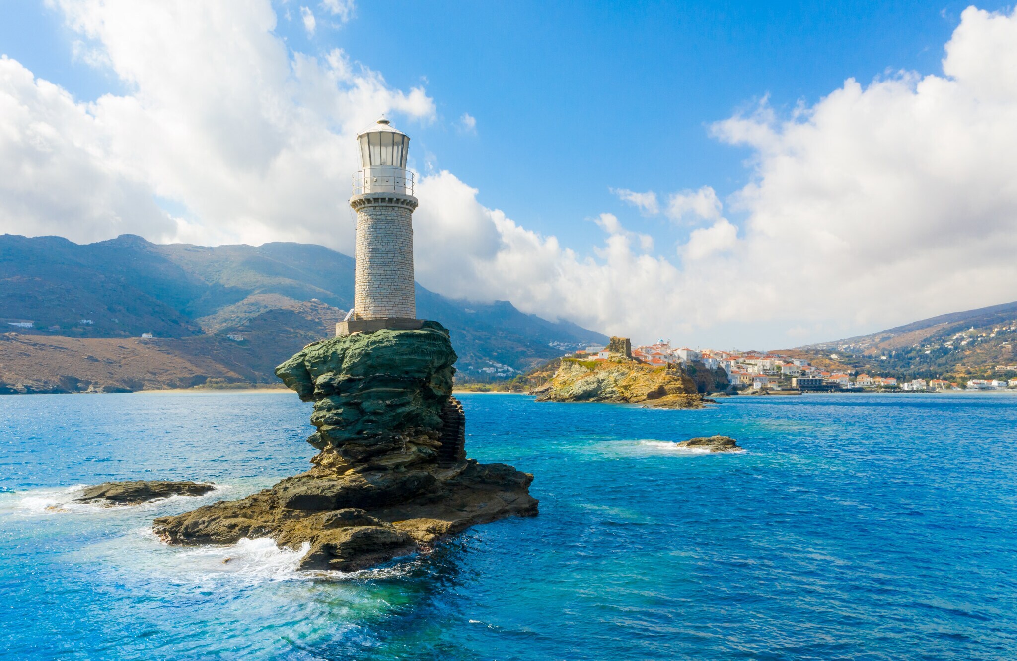 Weißer Leuchtturm auf einem kleinen Felsen im Meer vor einer Küste.
