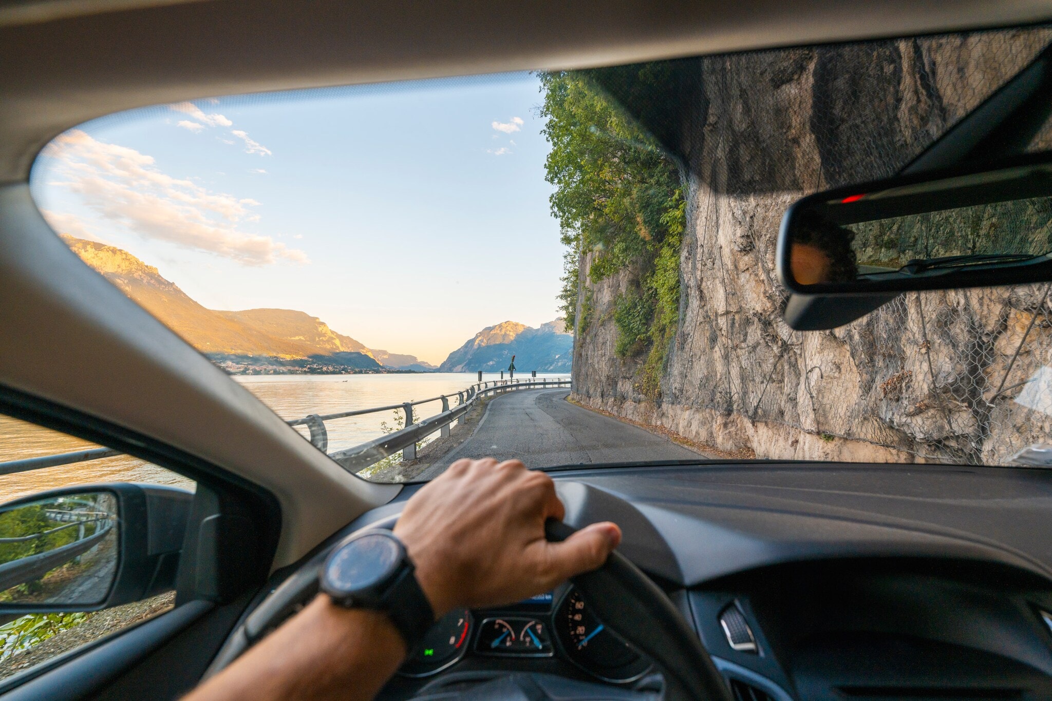 Blick auf eine Küstenstraße entlang einer Felswand durch die Windschutzscheibe eines Autos, im Vordergrund eine Hand am Lenkrad. Blick auf eine Küstenstraße entlang einer Felswand durch die Windschutzscheibe eines Autos, im Vordergrund eine Hand am Lenkrad.