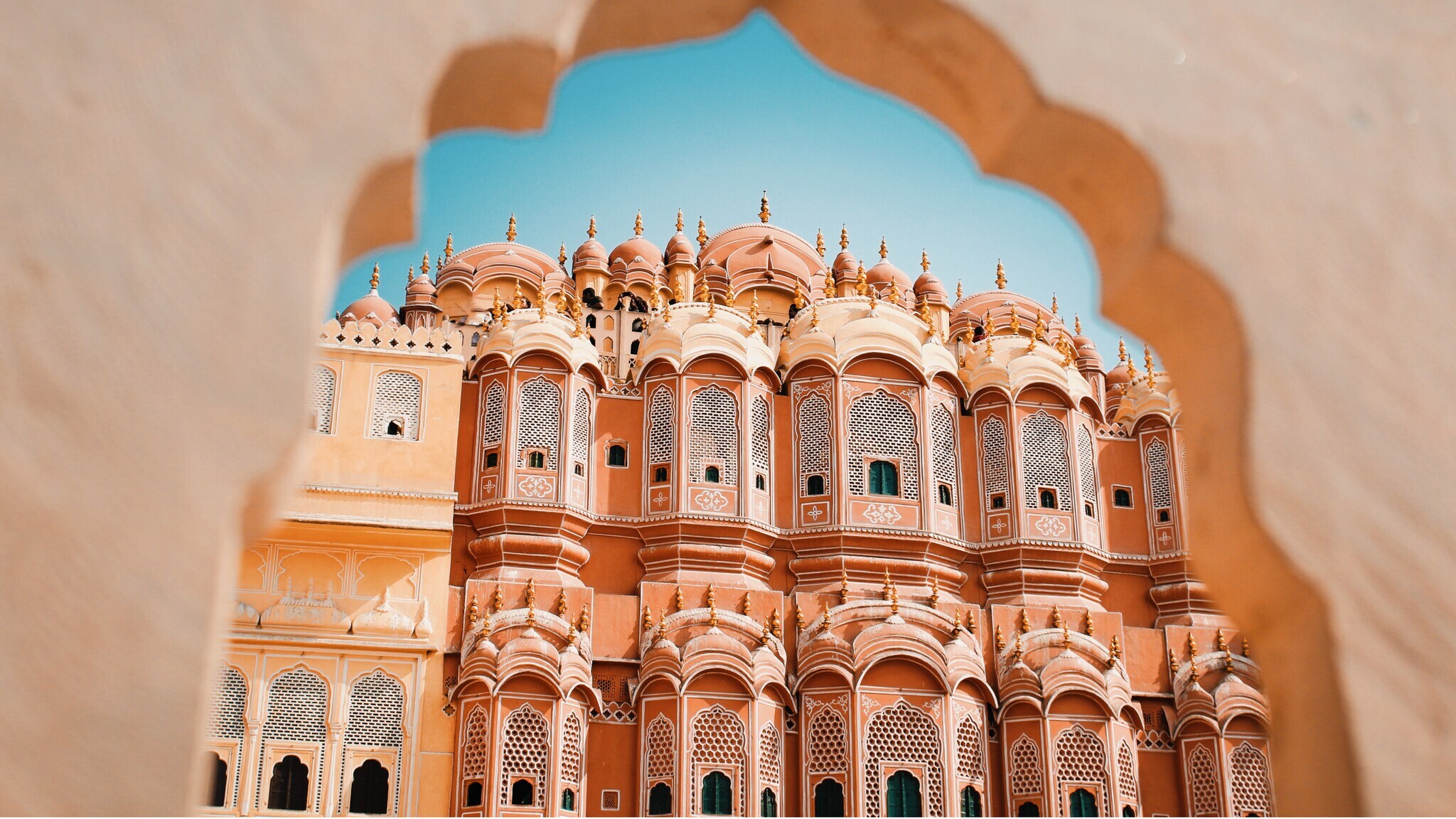 Blick durch ein kunstvoll geformtes Tor auf den Hawa Mahal, ein rosa Sandsteinpalast mit vielen Fenstern in Jaipur