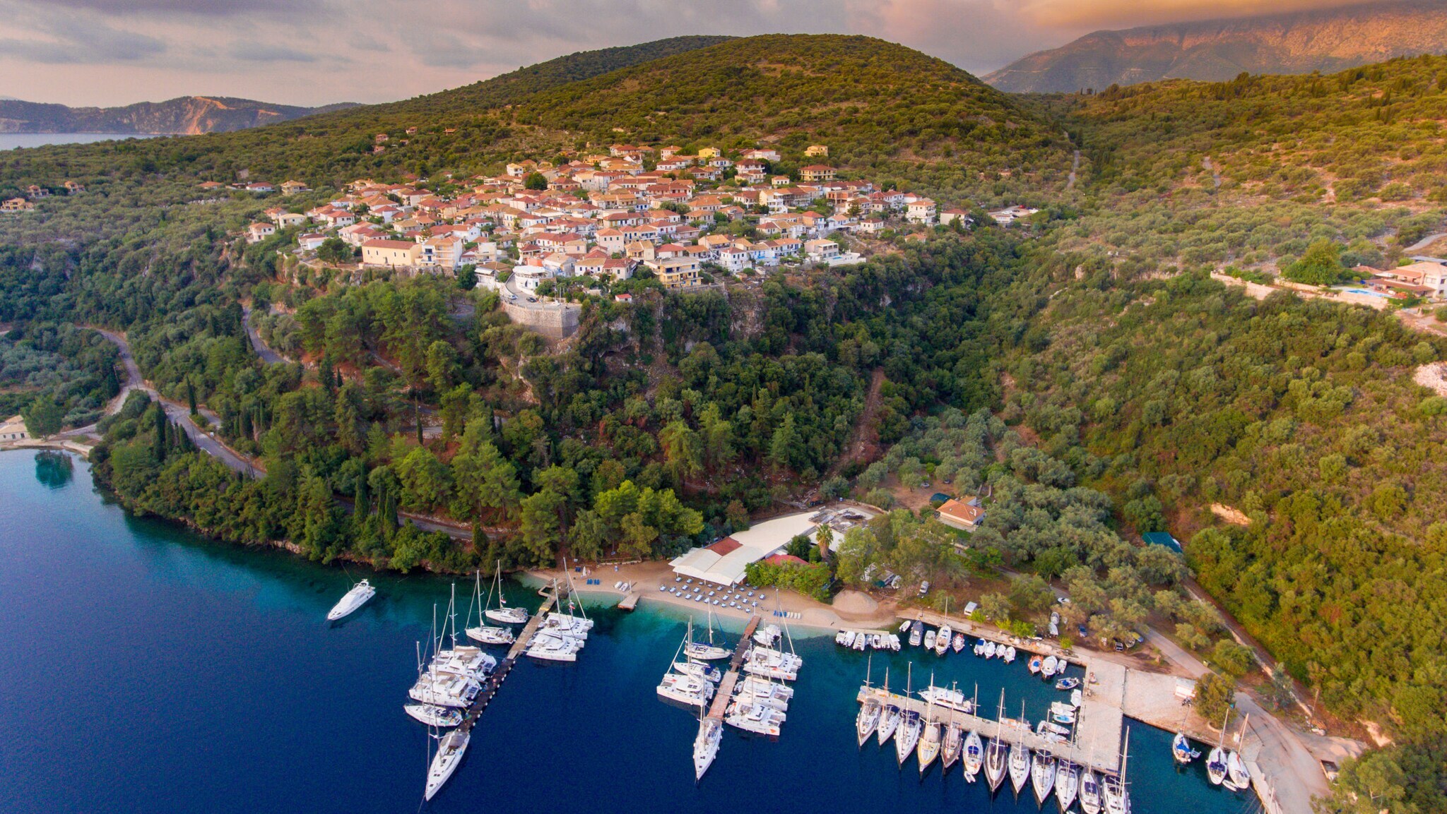 Luftaufnahme des Küstendorfes Meganisi mit Yachthafen, umgeben von bewaldeten Hügeln und ruhigem blauem Wasser Luftaufnahme des Küstendorfes Meganisi mit Yachthafen, umgeben von bewaldeten Hügeln und ruhigem blauem Wasser
