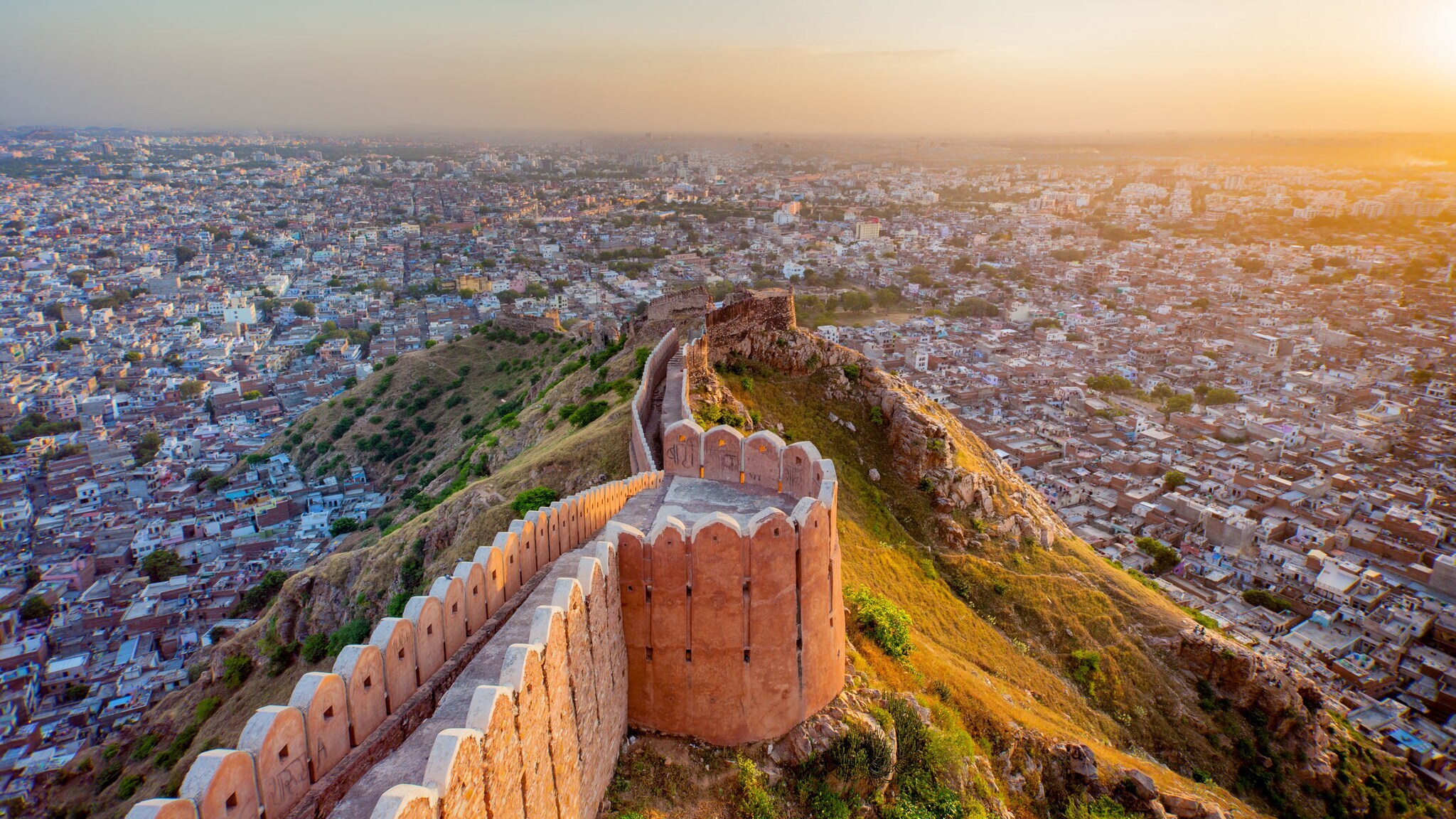 Luftaufnahme der historischen Stadtmauer von Jaipur, die sich über grüne Hügel erstreckt, mit der Stadt im Hintergrund bei Sonnenuntergang