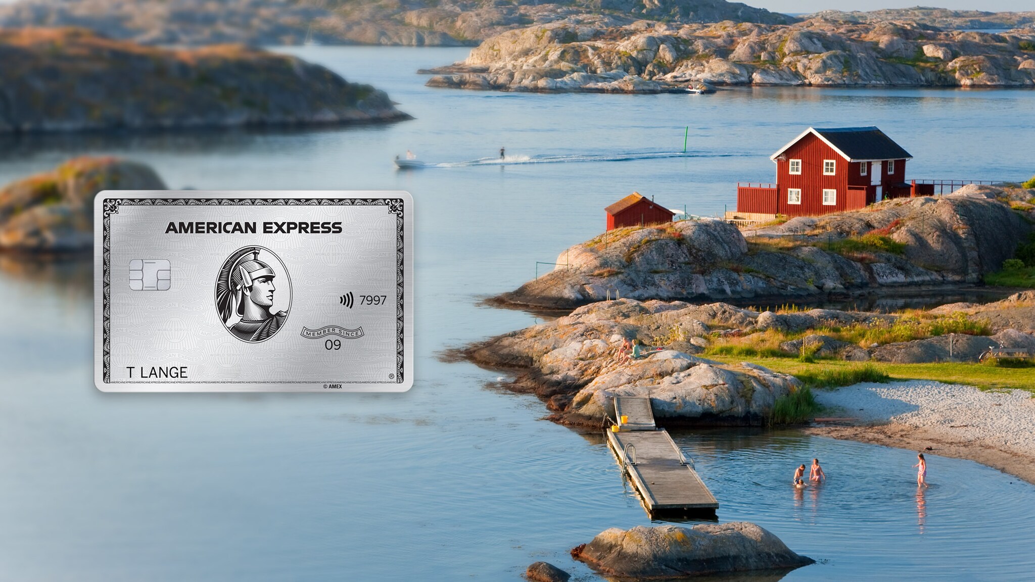 Schärenlandschaft mit Personen im Wasser vor einem roten Holzhaus im schwedischen Stil auf einem Felsen, im Vordergrund eine silberne Kreditkarte von American Express als Freisteller. Schärenlandschaft mit Personen im Wasser vor einem roten Holzhaus im schwedischen Stil auf einem Felsen, im Vordergrund eine silberne Kreditkarte von American Express als Freisteller.