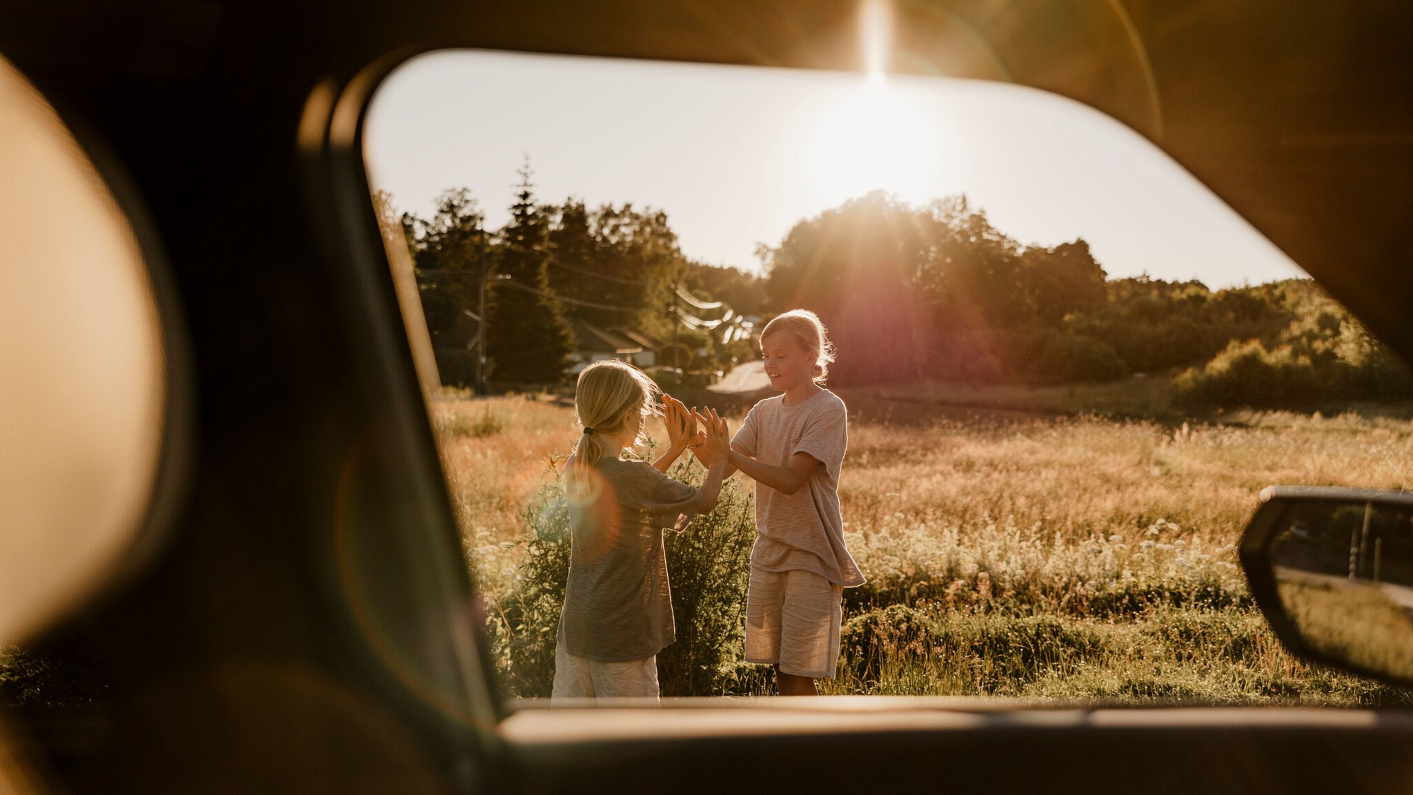Blick durch ein geöffnetes Autofenster auf zwei spielende Mädchen auf einer Wiese im Sonnenschein.