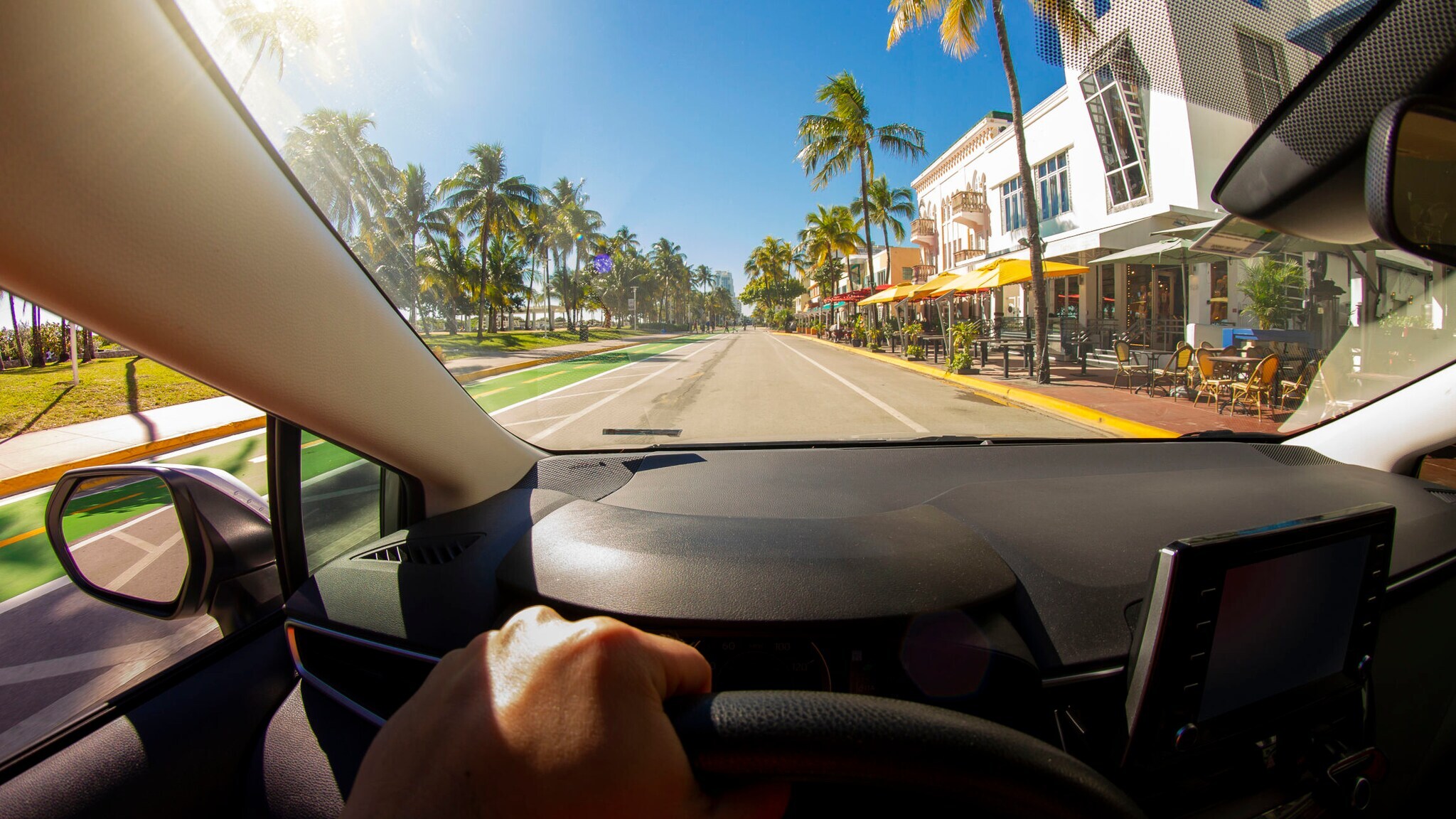 Aufnahme vom Fahrersitz eines Autos mit einer Hand am Lenkrad, das auf einer palmengesäumten Straße fährt Aufnahme vom Fahrersitz eines Autos mit einer Hand am Lenkrad, das auf einer palmengesäumten Straße fährt
