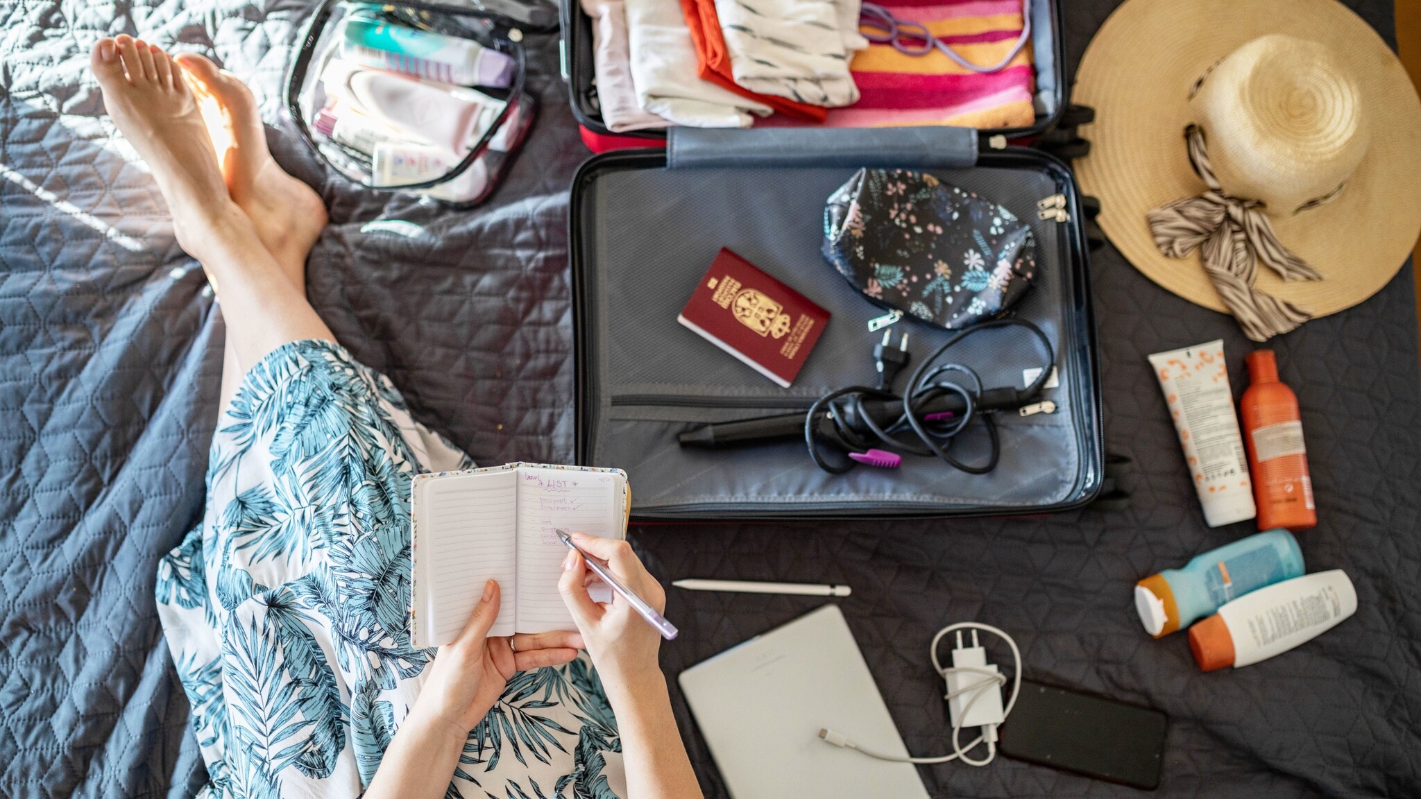 Aufsicht eines Koffers und ausgebreiteten Reiseutensilien auf einem Bett, daneben Beine und Hände einer Frau, die in ein Notizheft schreibt. Aufsicht eines Koffers und ausgebreiteten Reiseutensilien auf einem Bett, daneben Beine und Hände einer Frau, die in ein Notizheft schreibt.