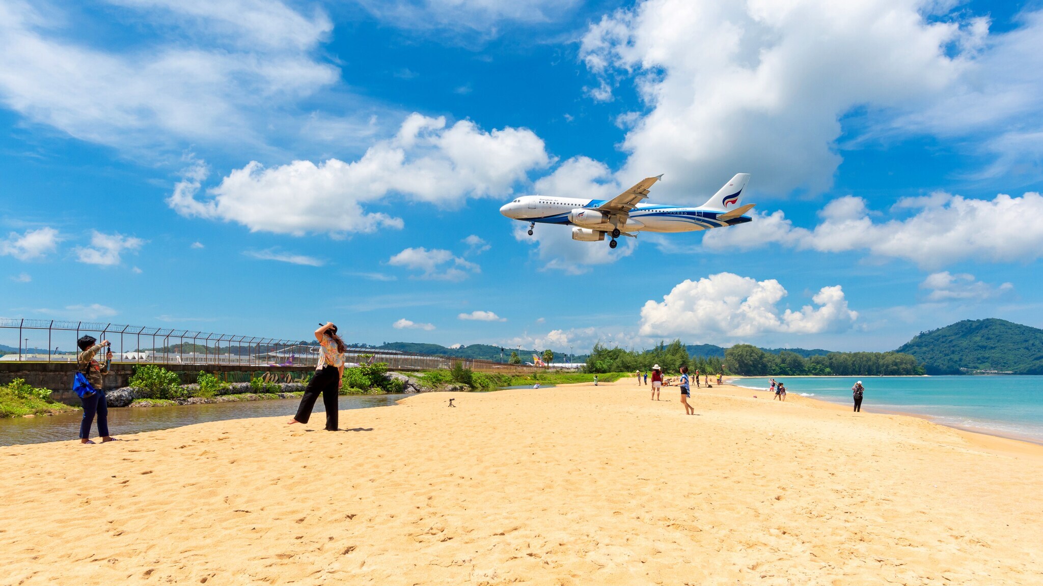 Personen auf einem Sandstrand an einem eingezäunten Gebiet, über dem ein Flugzeug im Landeanflug fliegt.