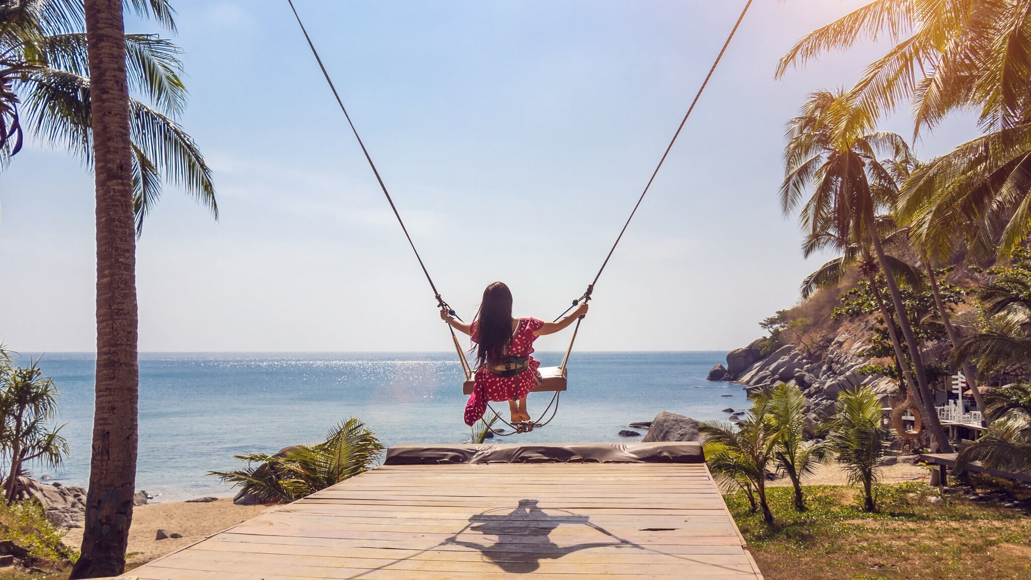 Rückansicht einer Frau im roten Kleid auf einer Schaukel an einem Strand, umgeben von Palmen. Rückansicht einer Frau im roten Kleid auf einer Schaukel an einem Strand, umgeben von Palmen.