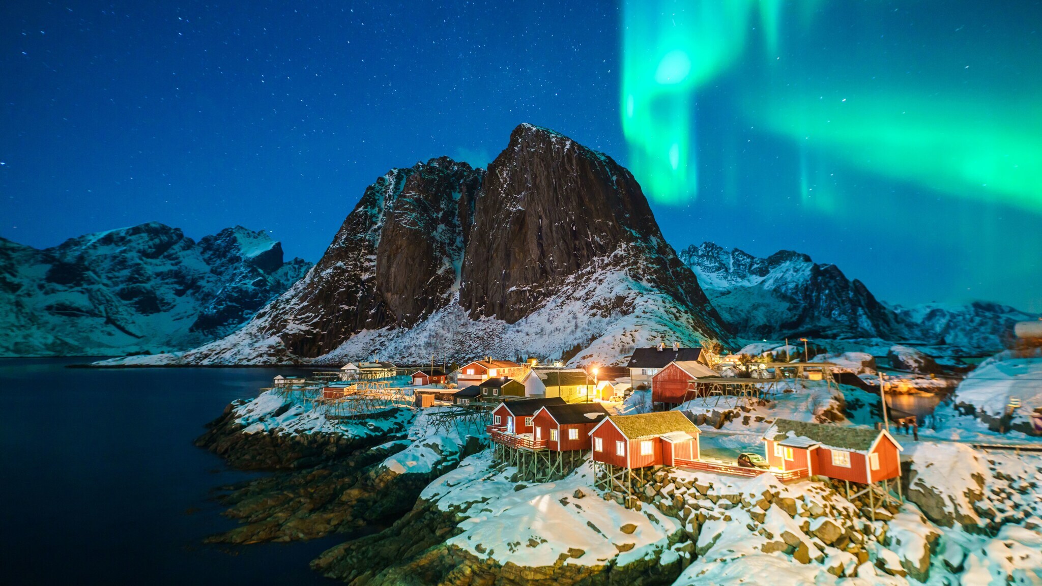 Siedlung mit bunten Holzhäusern in schneebedeckter Felslandschaft unter Polarlichtern bei Nacht.