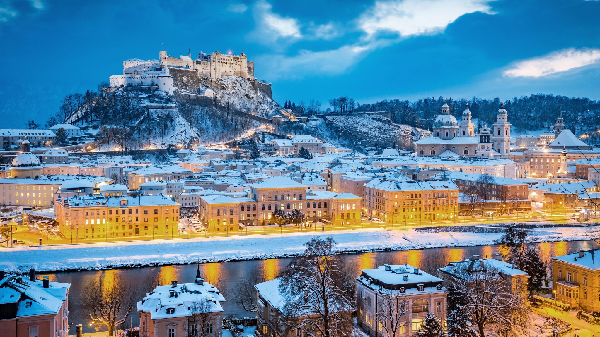 Schneebedecktes Stadtpanorama von Salzburg mit beleuchteten Straßen bei Dunkelheit.