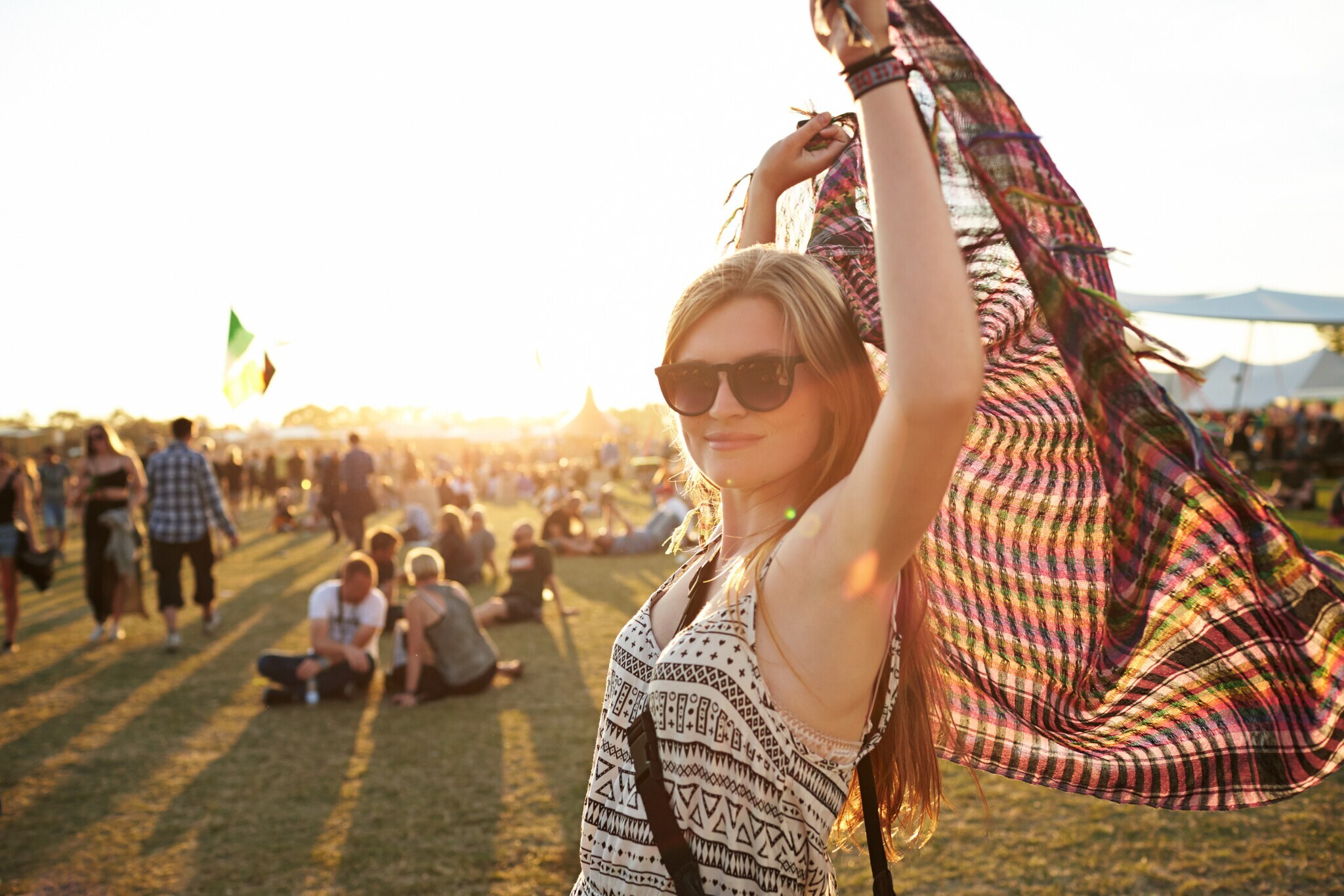 Eine Frau mit Sonnenbrille steht auf einer Wiese, auf der Leute sitzen, und hält ein Tuch über ihren Kopf.